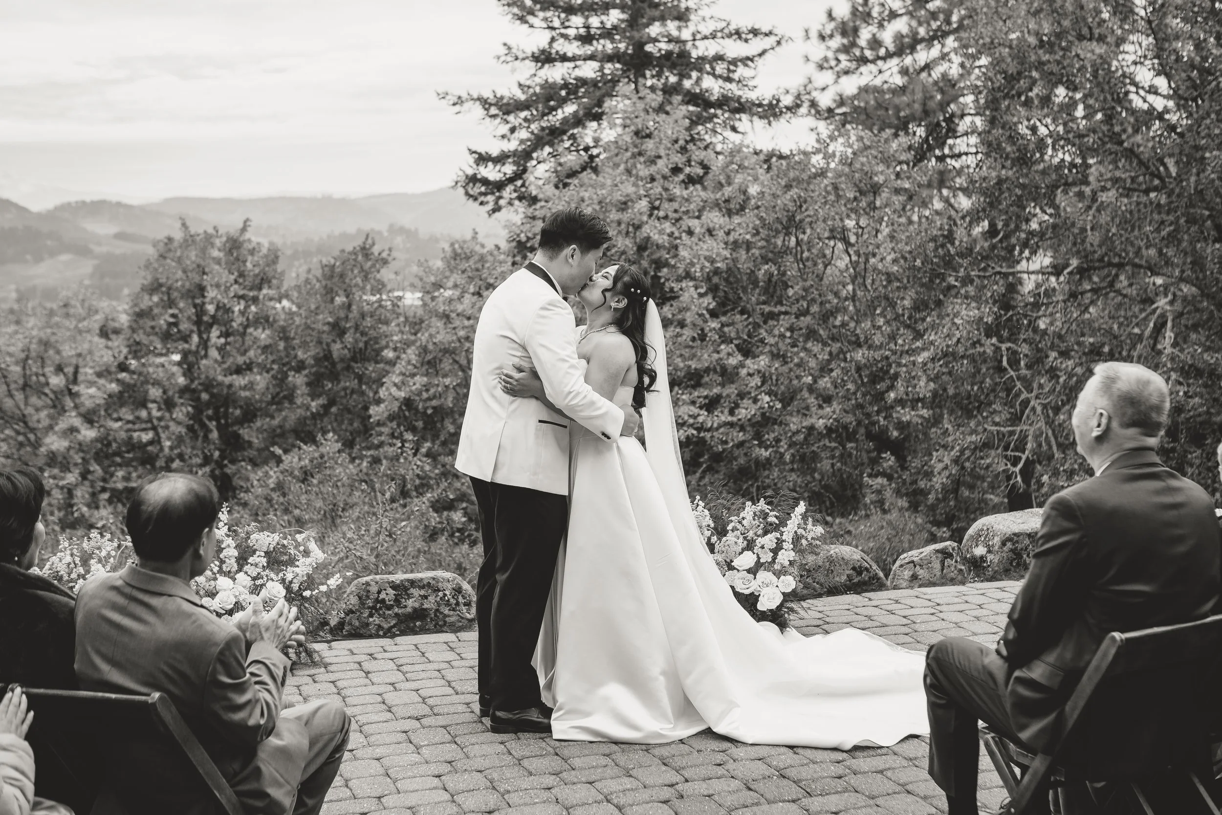 A black and white photo of a bride and groom sharing a kiss during their outdoor wedding ceremony, with guests seated around them and trees in the background.