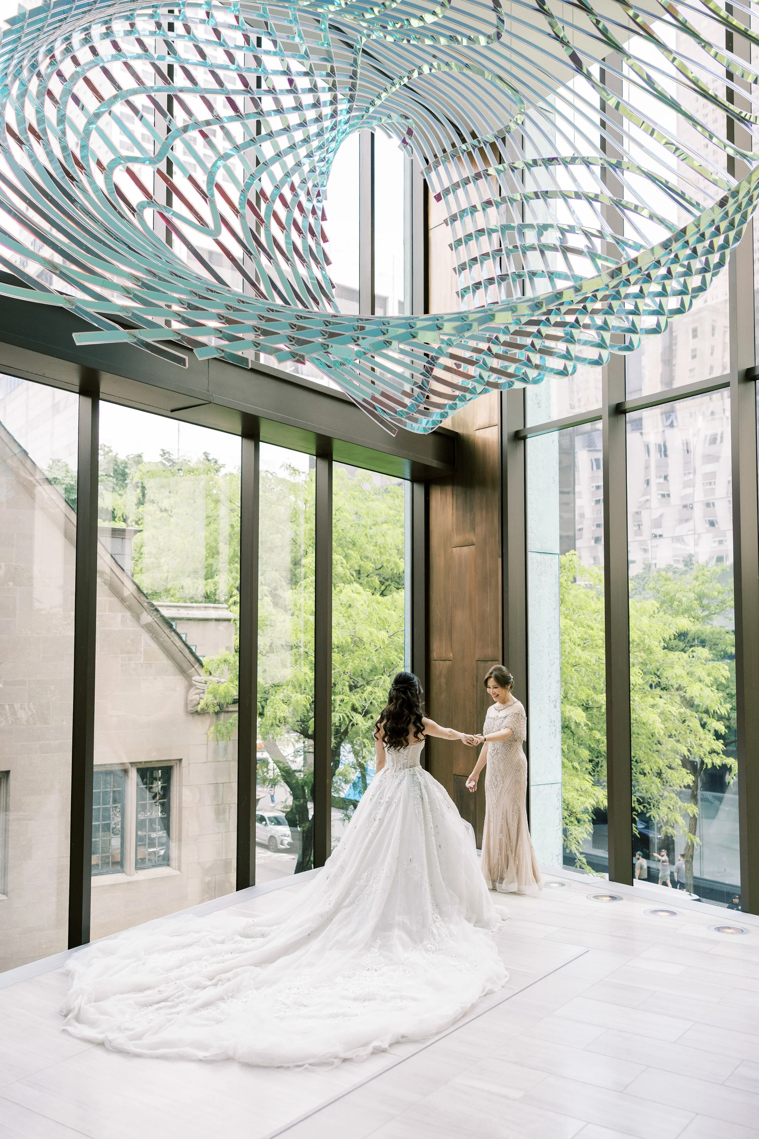 Two women dancing during a wedding ceremony inside a modern glass building with a decorative metallic sculpture overhead, and city buildings and trees visible through large windows outside.