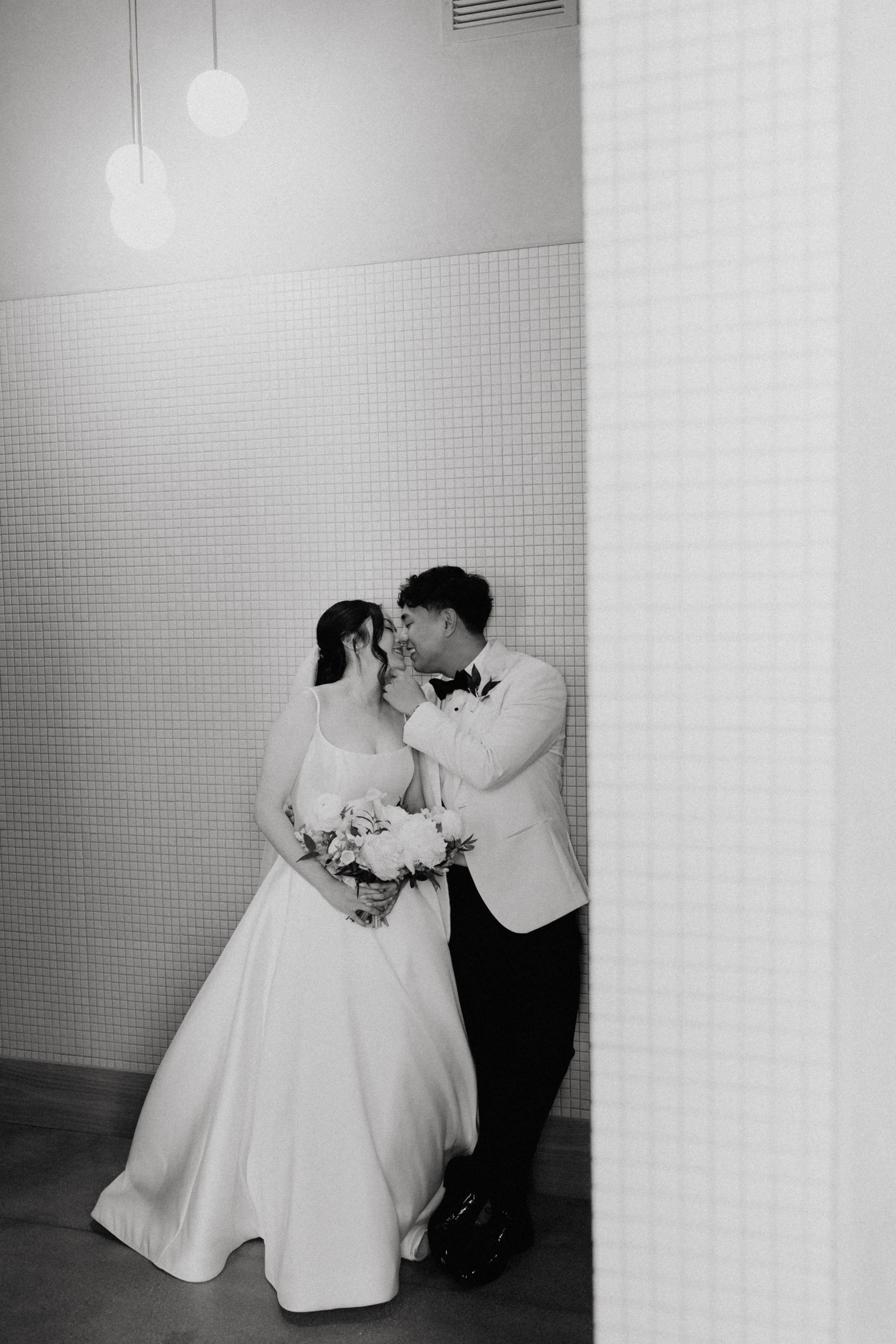 A couple dressed in wedding attire embracing each other and touching foreheads, standing against a textured wall. The bride holds a bouquet of flowers, and they are smiling with closed eyes.