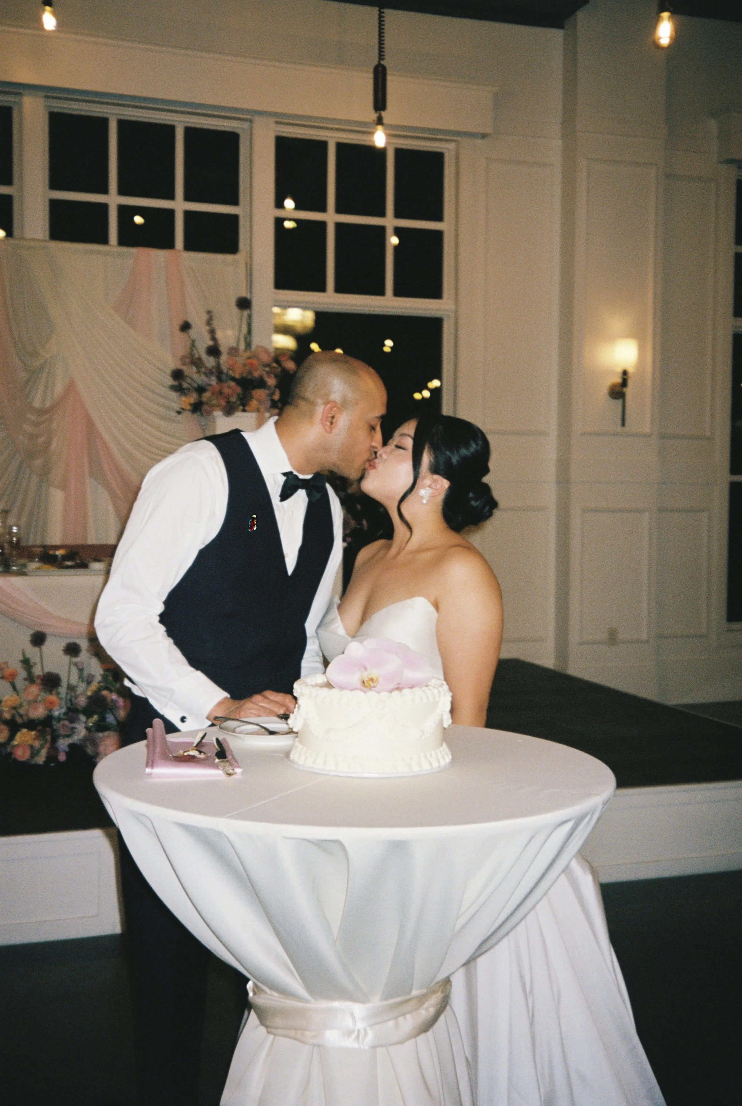 A bride and groom share a kiss at their wedding reception. The bride is wearing a strapless white gown and holding a bouquet of flowers. The groom is dressed in a black vest, white shirt, and black bow tie. A wedding cake is on the table in front of 