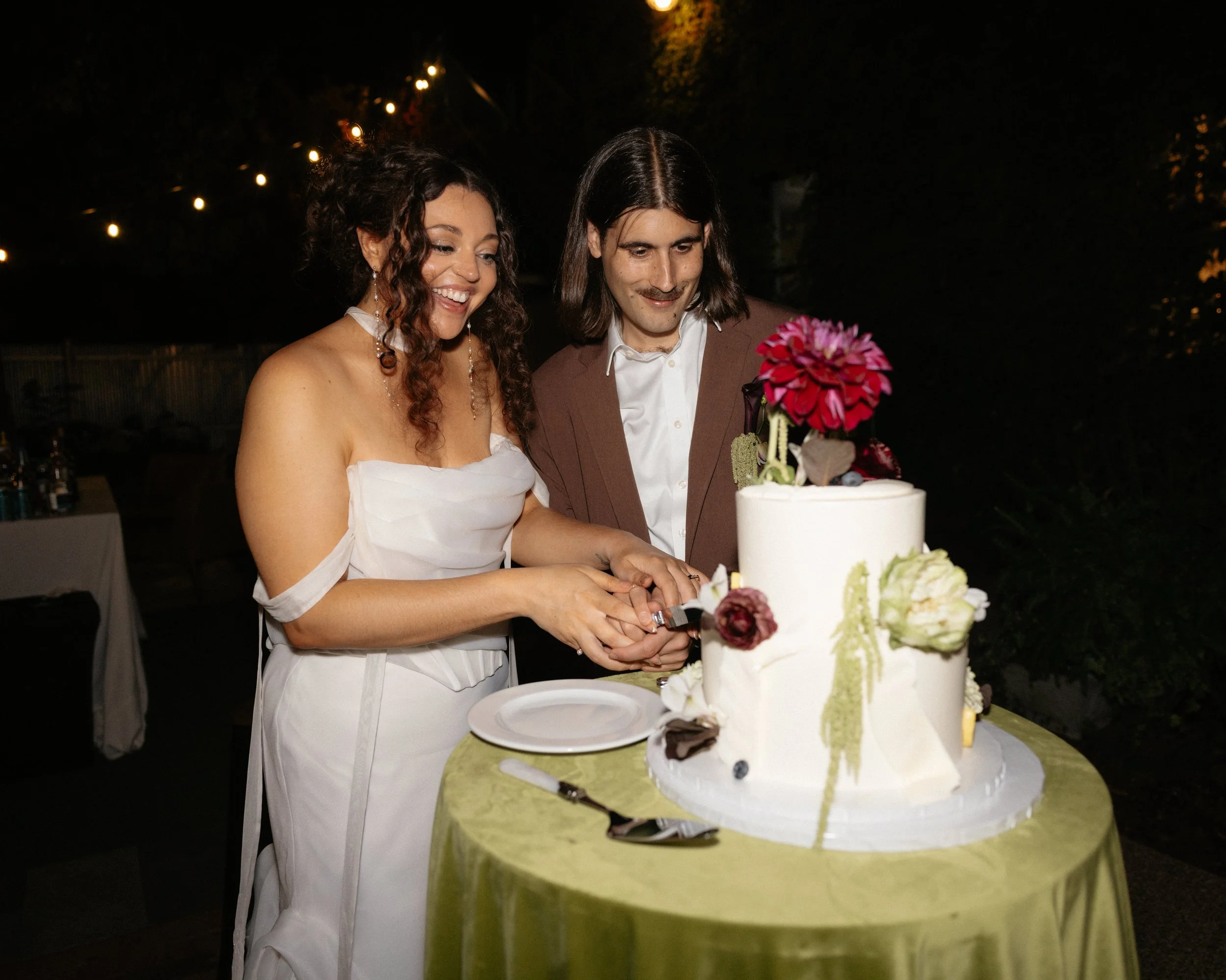 A bride and groom cutting a wedding cake outdoors at night, surrounded by string lights.