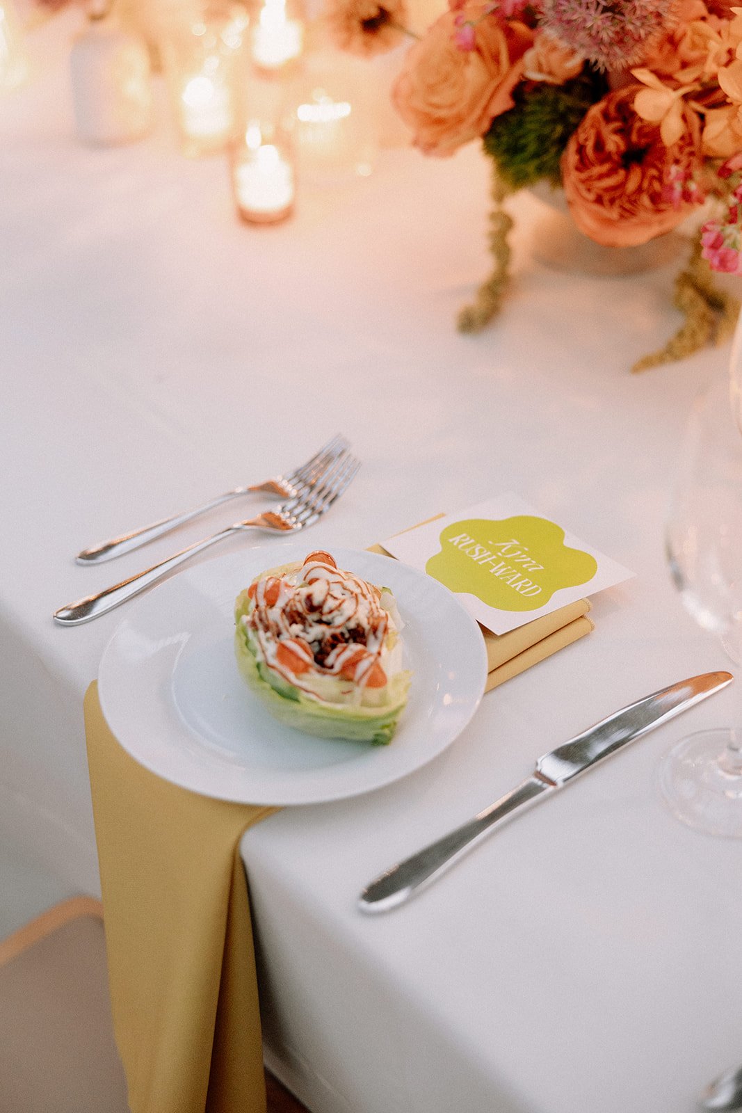 A dessert on a white plate with cream, strawberries, and drizzle, placed on a beige napkin on a white tablecloth. Silverware and a bouquet of pink and peach flowers are visible in the background, with candles creating a warm glow. A place card reads 