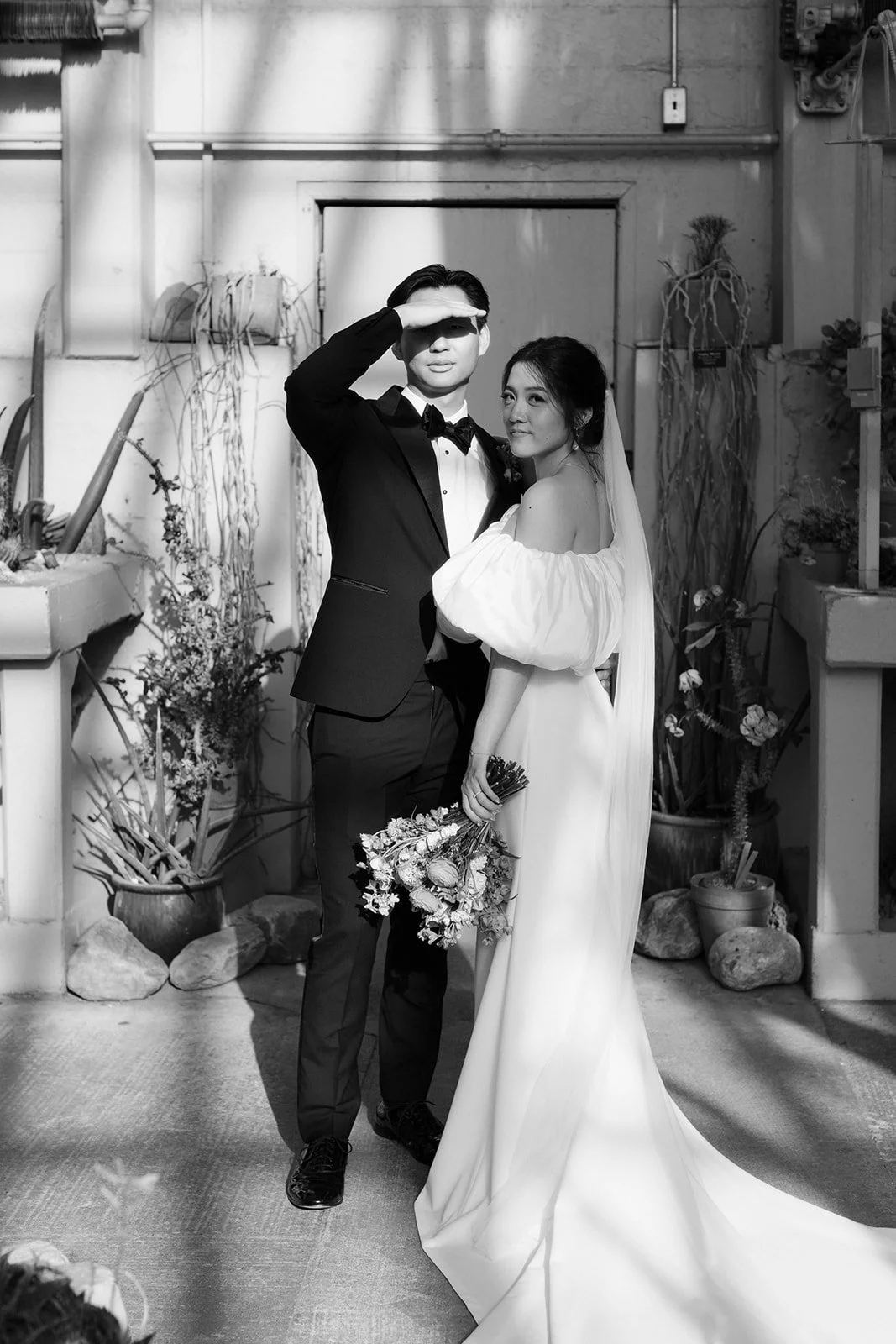 Black and white photo of a bride and groom at their wedding, standing close together with the groom saluting and the bride holding a bouquet, in an indoor garden setting with plants and stones.