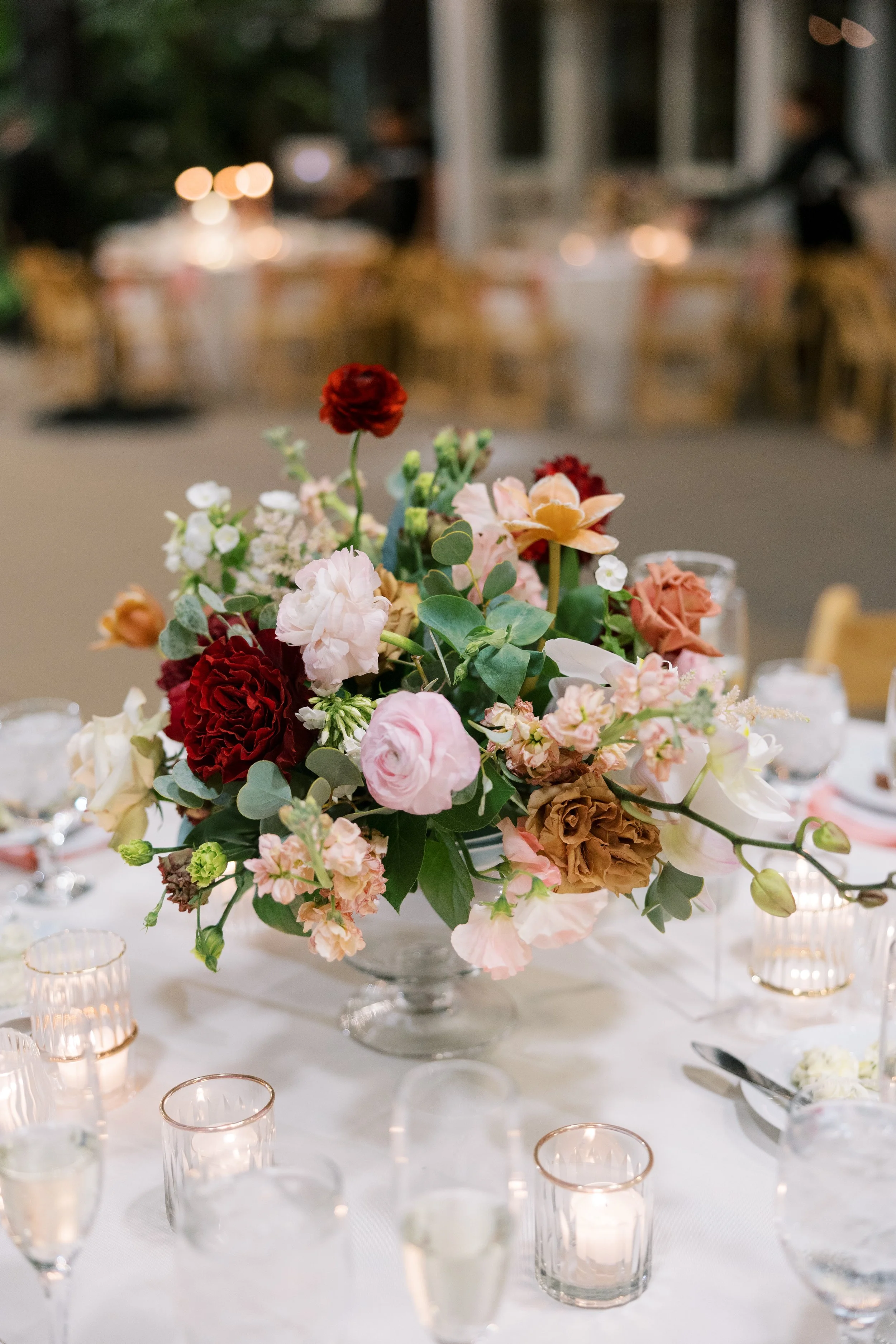 A floral centerpiece with pink, red, and peach flowers on a table surrounded by votive candles in glass holders at an elegant event.