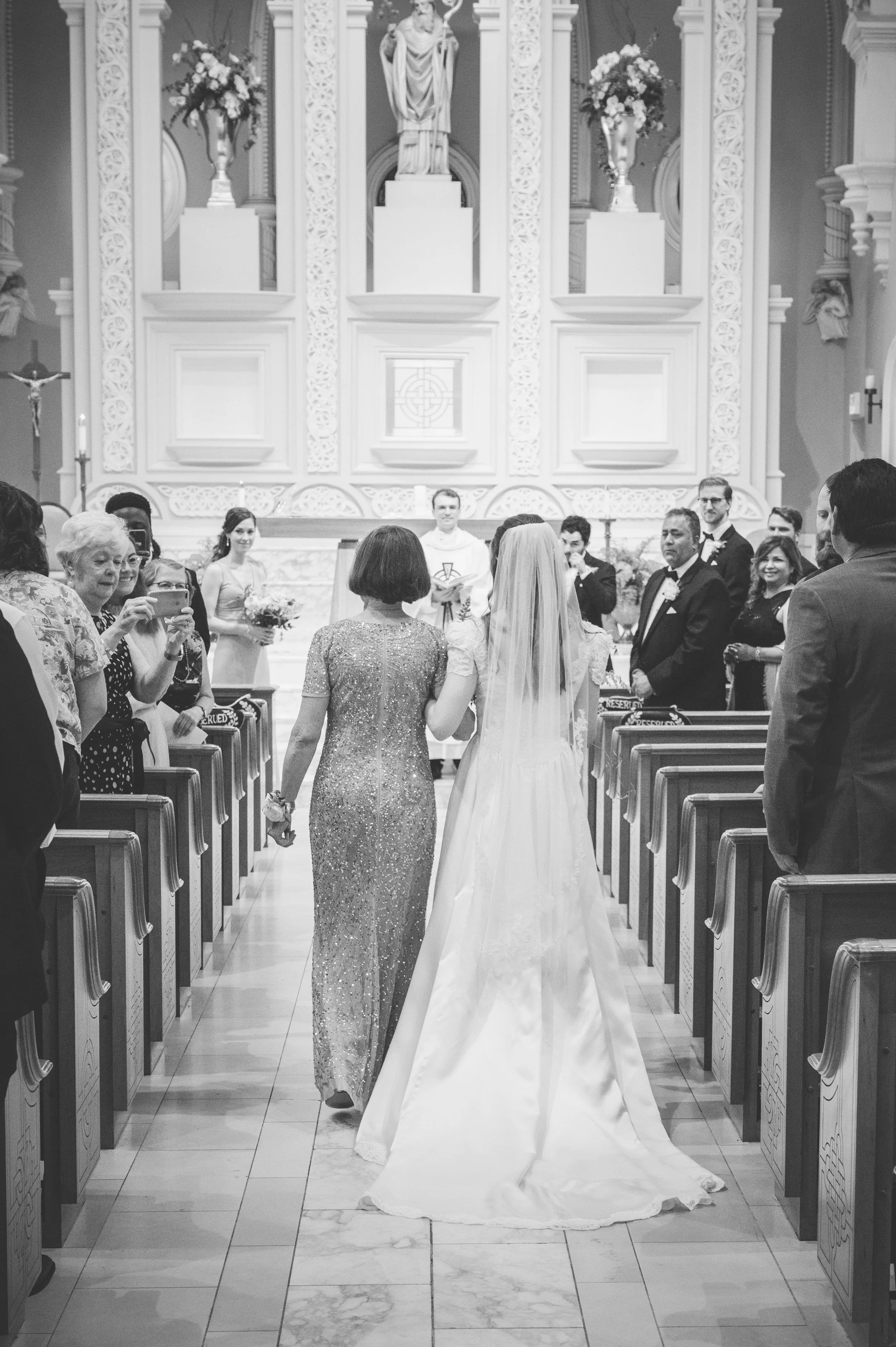 A wedding ceremony inside a church, with a bride and another woman holding hands and walking down the aisle, surrounded by seated guests and a priest in the background.