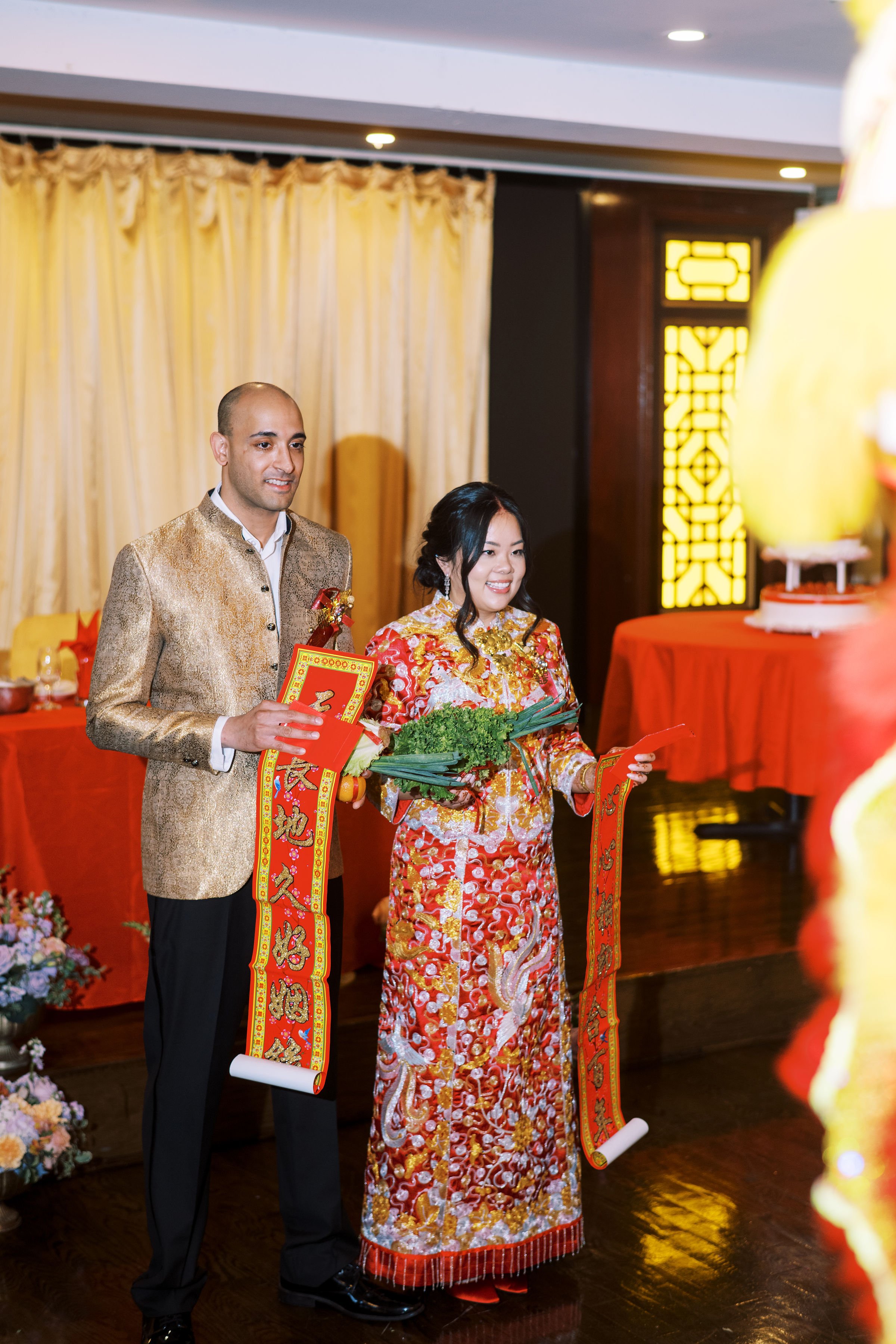 A man and woman dressed in traditional Chinese wedding attire holding red envelopes and a bouquet of flowers in a decorated reception area with gold curtains and red tables.