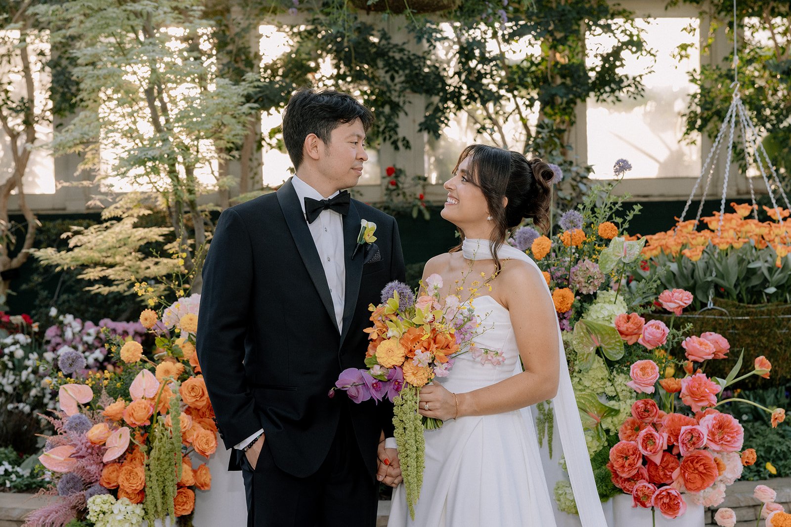 A bride and groom stand close, holding hands and smiling at each other, in a garden setting with vibrant orange, pink, and purple flowers behind them.