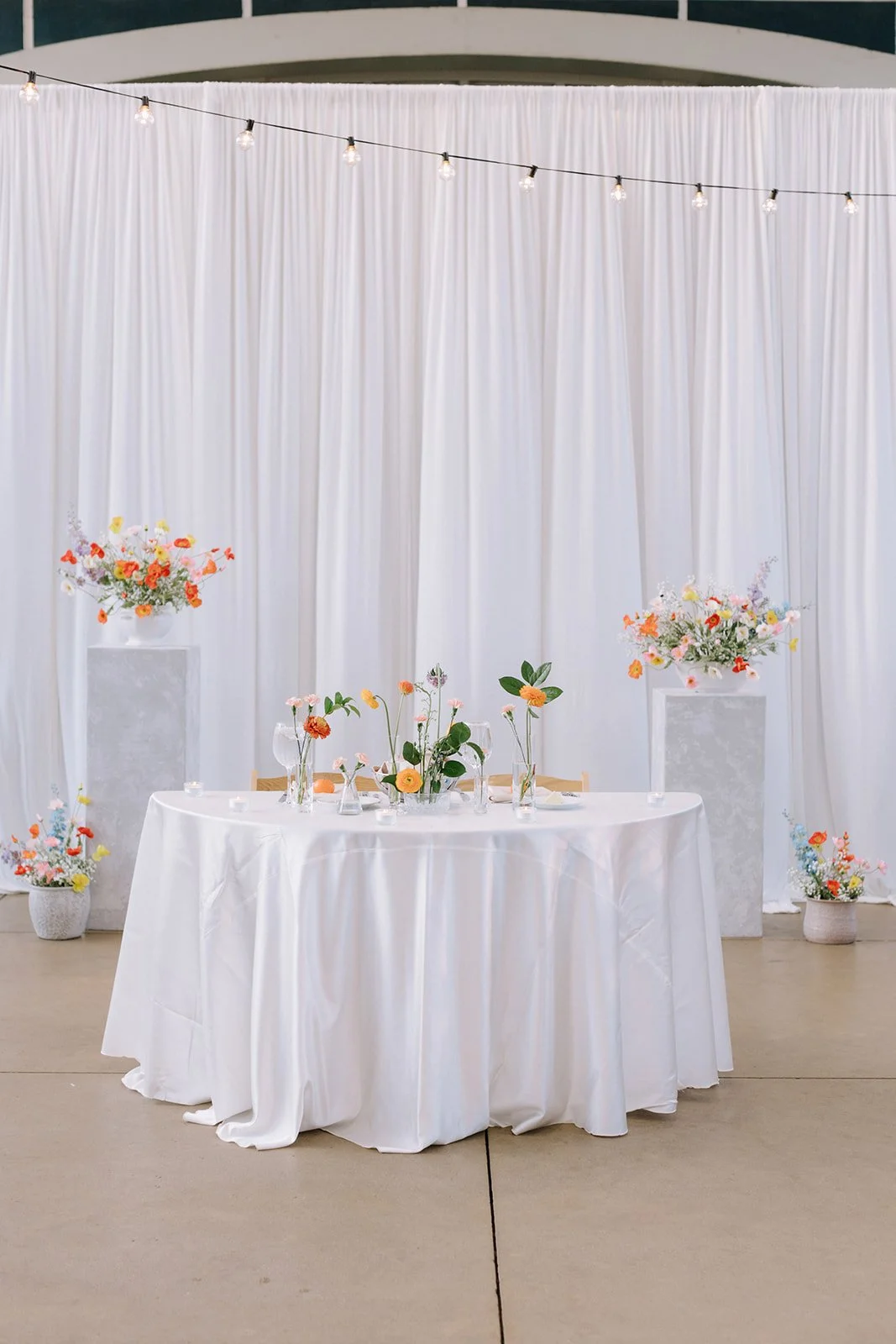 Elegant wedding or event table setup with a white tablecloth, floral centerpieces, and pastel-colored flowers in vases. The backdrop consists of white curtains and tall vases with flower arrangements, with string lights hanging above.