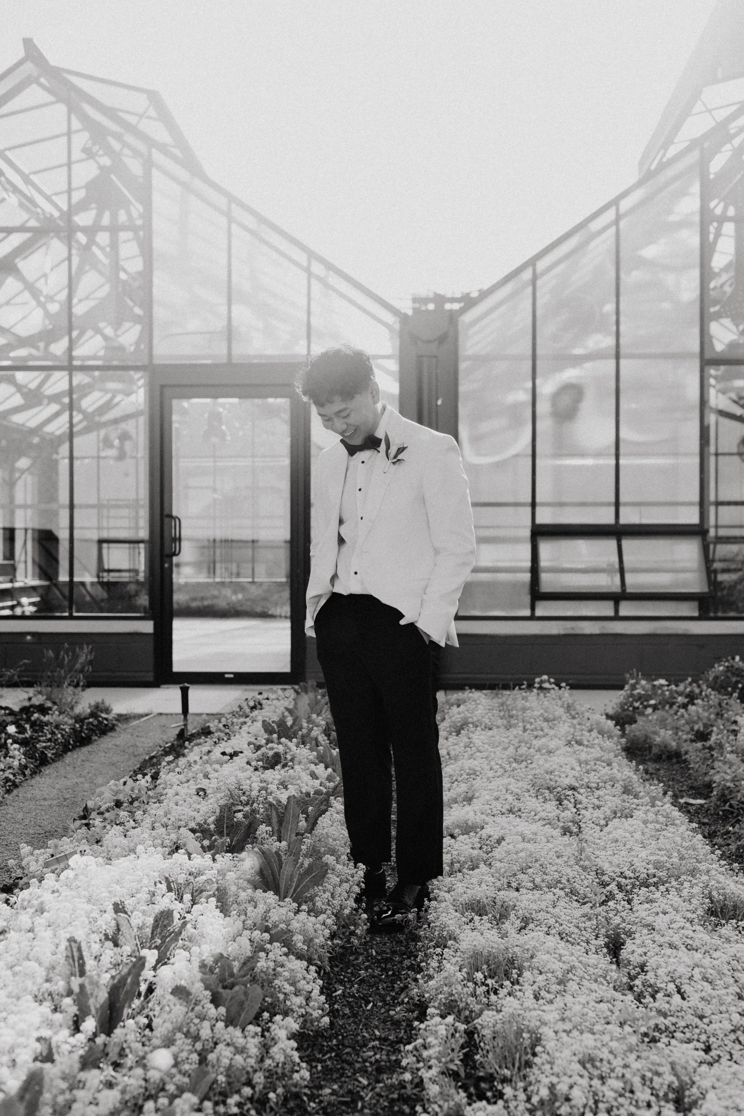 A young man in a tuxedo stands in a garden with flowers, smiling while looking down, with greenhouse structures in the background.