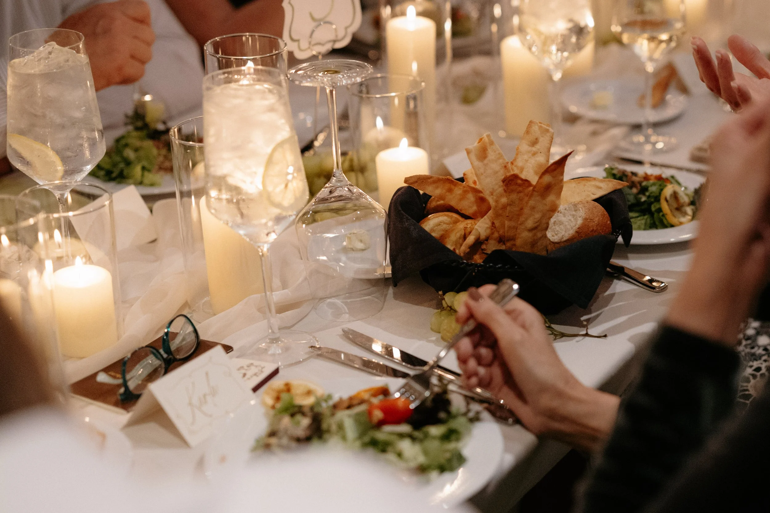 A table set for a formal dinner with candles, glasses of water, bread, salads, and other dishes. People are participating in the meal, some holding utensils.