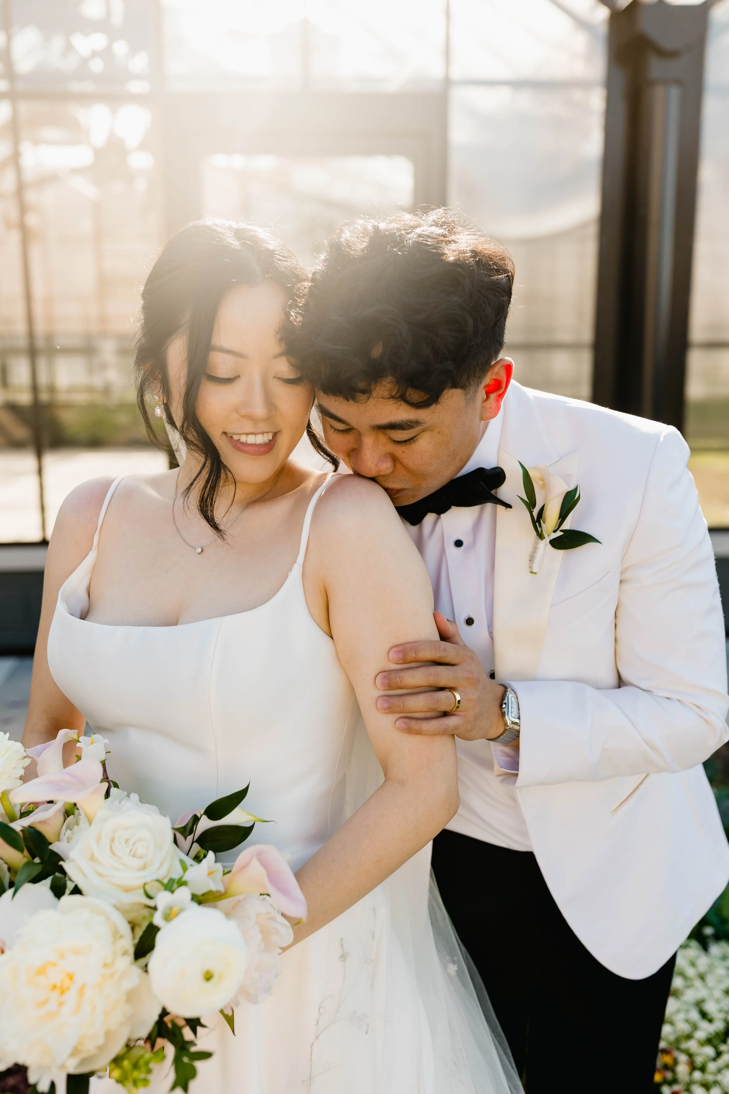 A newlywed couple, with the groom kissing the bride's shoulder, standing outdoors near a glass structure with sunlight shining in the background.