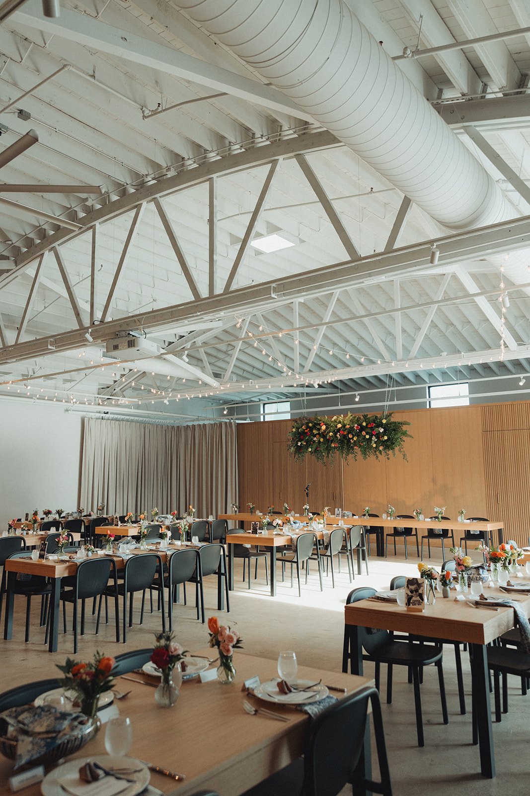 Interior of a decorated event space with tables set for a celebration, featuring elegant floral centerpieces, hanging string lights, and a flower arrangement above the main stage area. The ceiling has exposed beams and ductwork, with natural light co