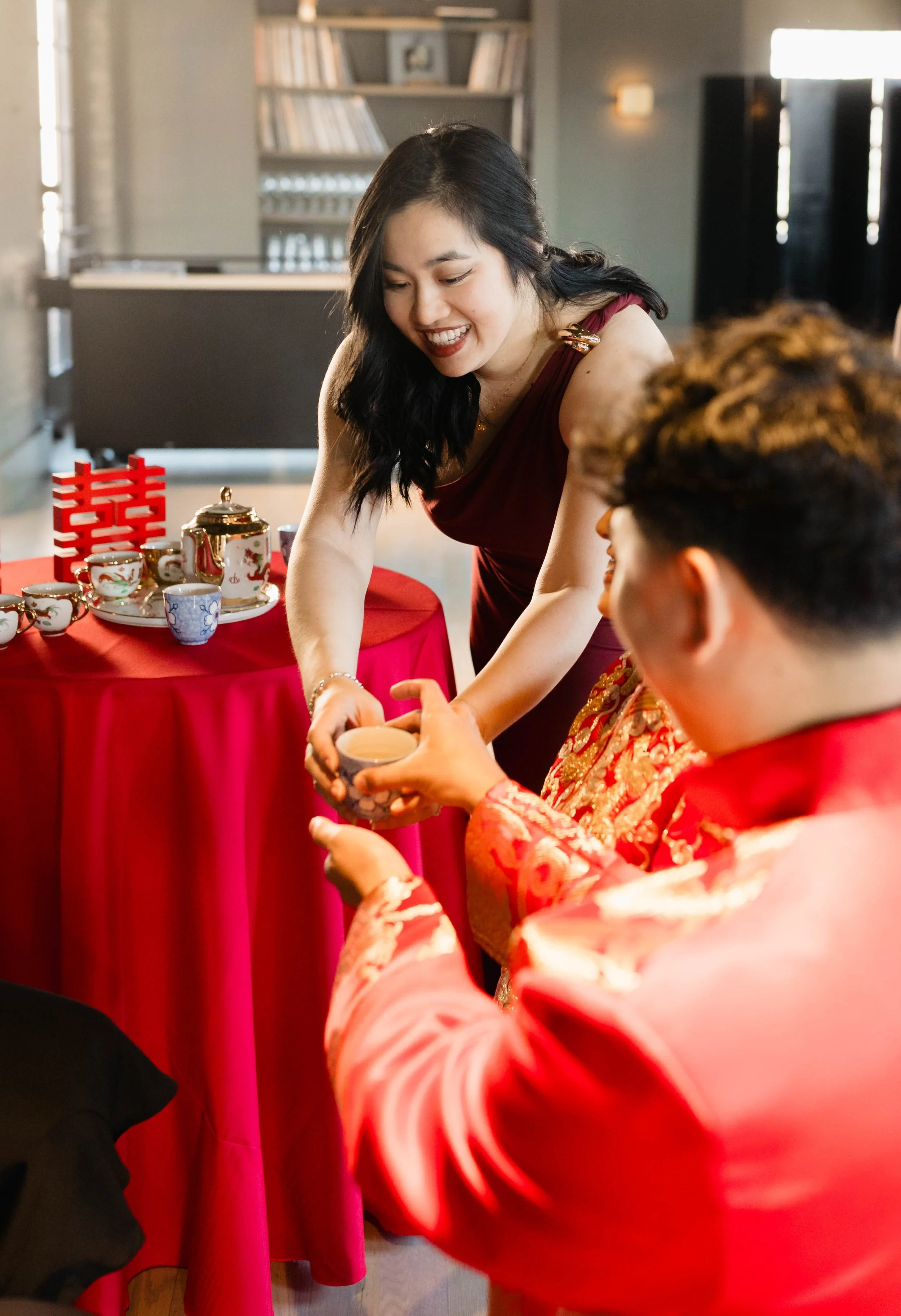 A woman in a maroon dress is presenting a tea cup to a man in a red and gold traditional Chinese outfit. There are tea cups and a teapot on a table with a red tablecloth, and a red decorative item on the table. They are indoors, in a room with bookshelves and a black door in the background.