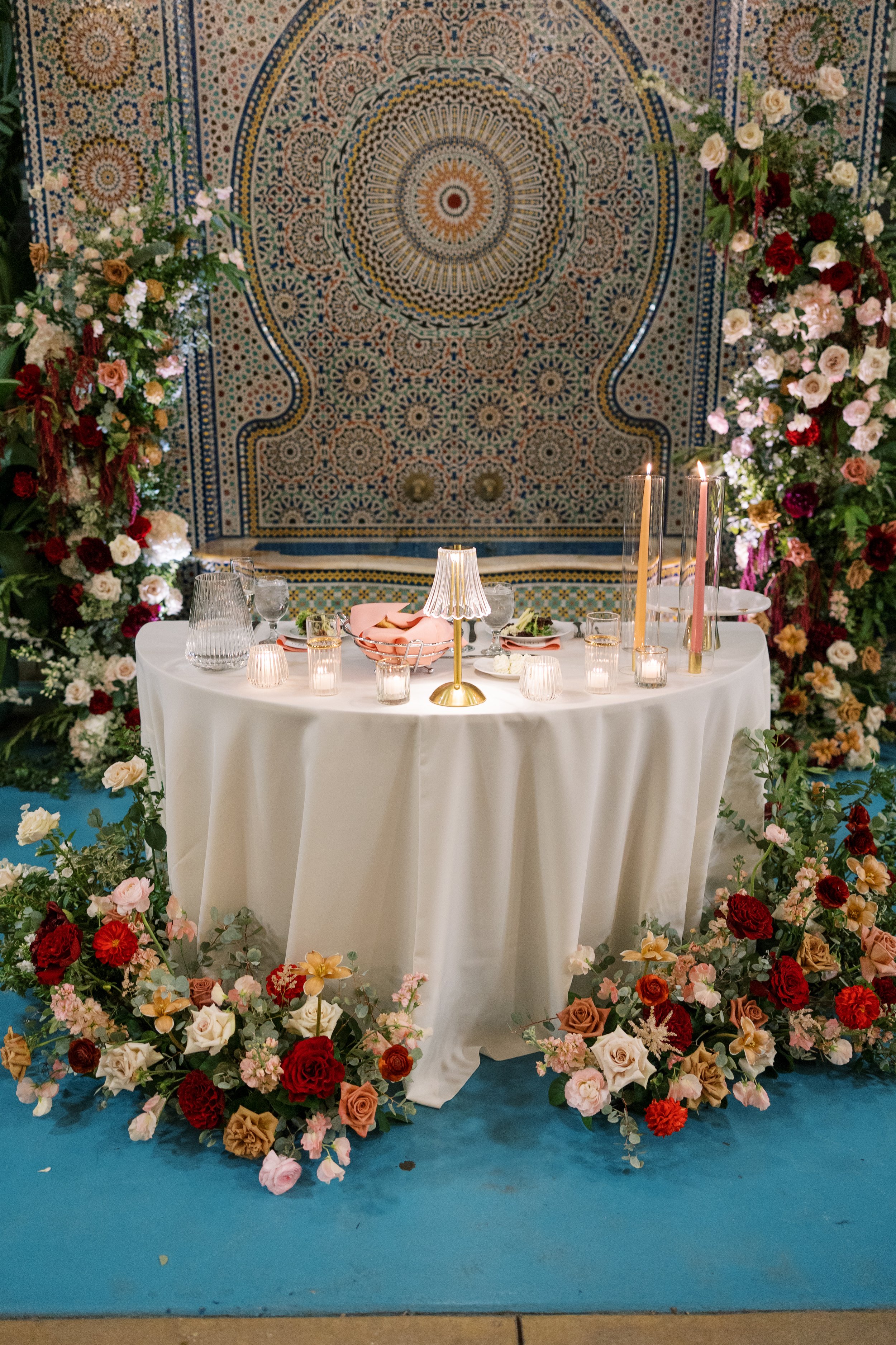 A decorated table with a white tablecloth, surrounded by colorful flowers, with candles and a pink cloth on it, in front of a large, ornate mosaic wall with a circular pattern.