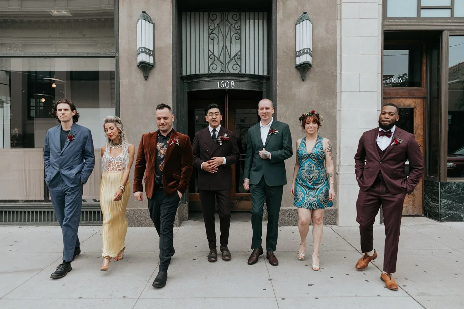 Group of eight diverse people walking on city sidewalk in front of building with address 1608, dressed in formal clothing, some with boutonnières and one with a floral crown, smiling and enjoying a sunny day.