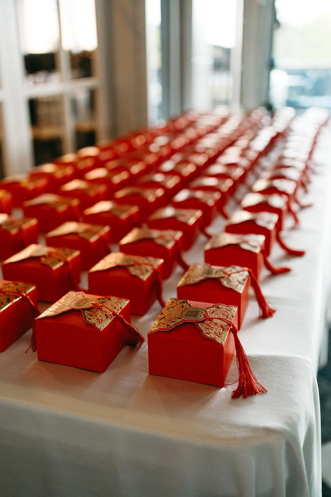 Multiple red gift boxes with gold-patterned lids and red tassels, arranged in rows on a white table near windows.