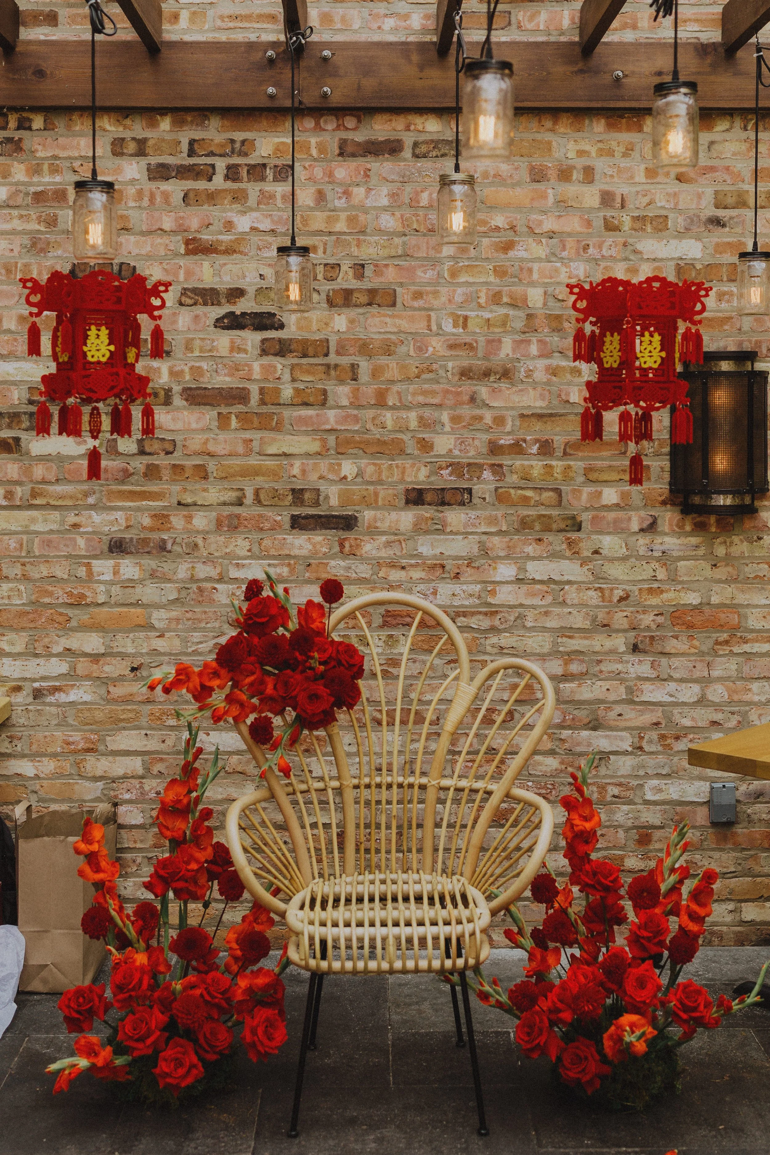A rattan chair in front of a brick wall with hanging industrial-style lights, two red Chinese lantern decorations, and a table with red and orange flowers in a vase.