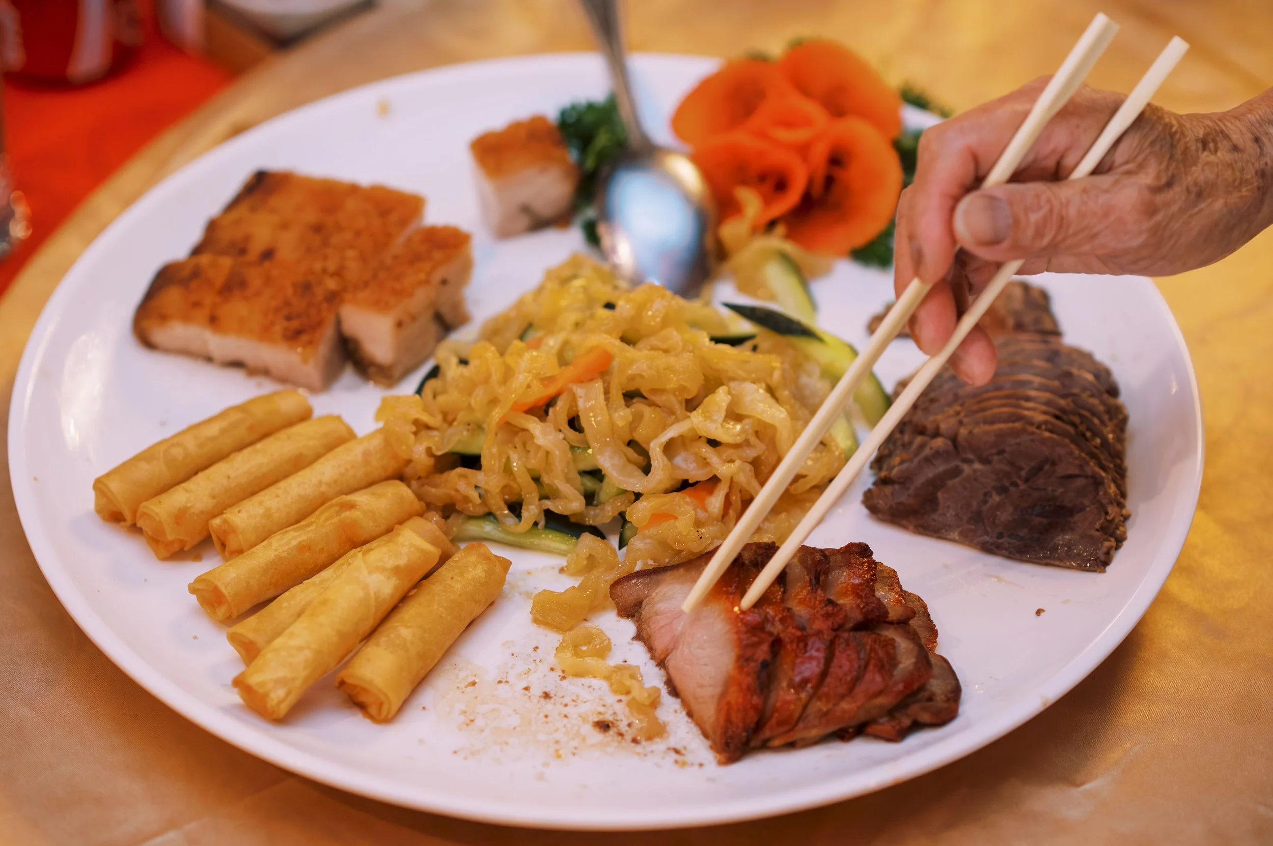A white plate filled with various Asian dishes. There are fried spring rolls, sliced grilled meat, a portion of stir-fried cabbage, and sliced grilled pork. The plate also has some sliced tomatoes and a few pieces of lettuce as garnish. A person's ha