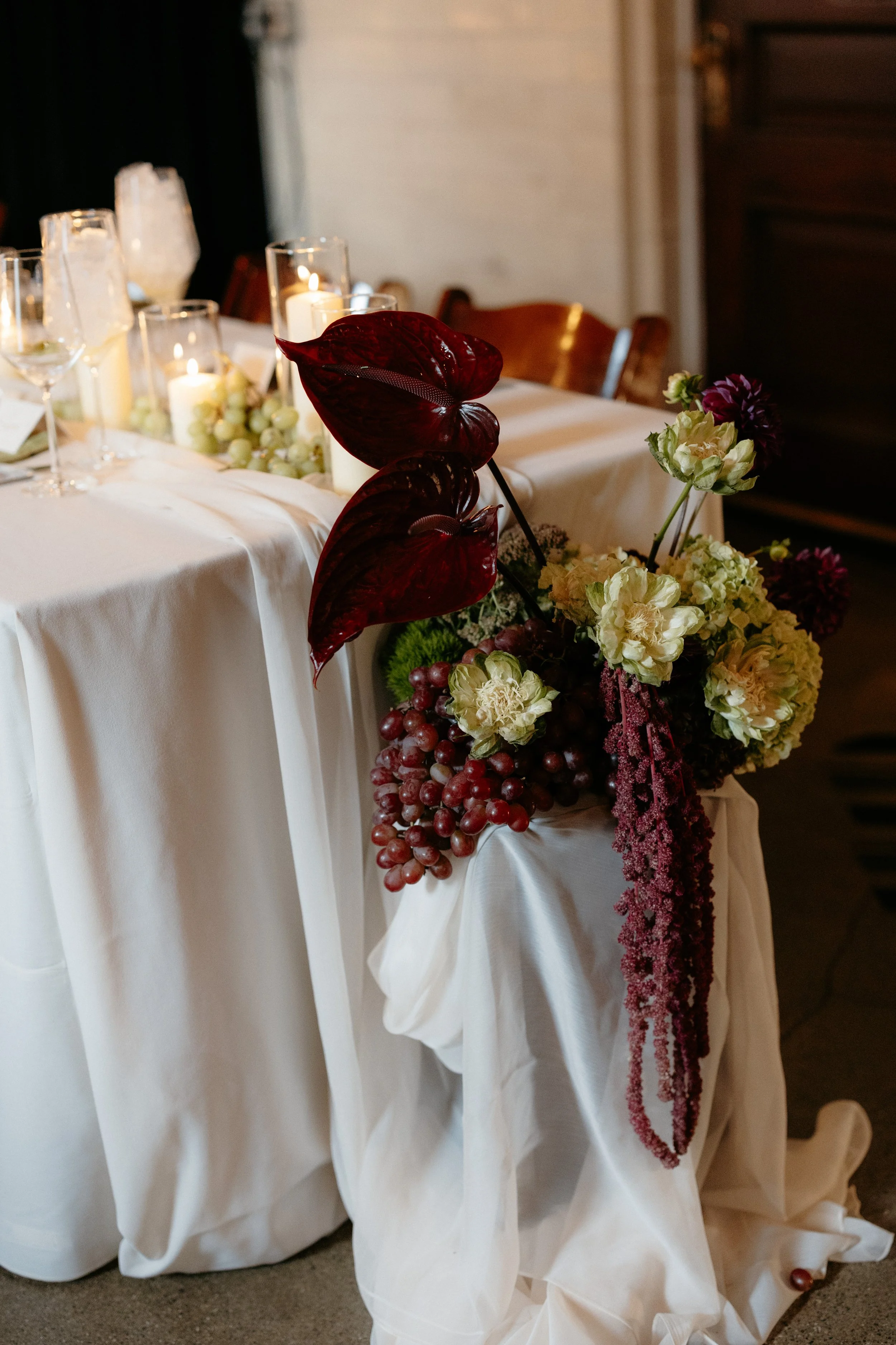 A table decorated with white cloth, candles, grape clusters, and an arrangement of flowers including dark red and white blossoms, with a large dark red leaf in the foreground.