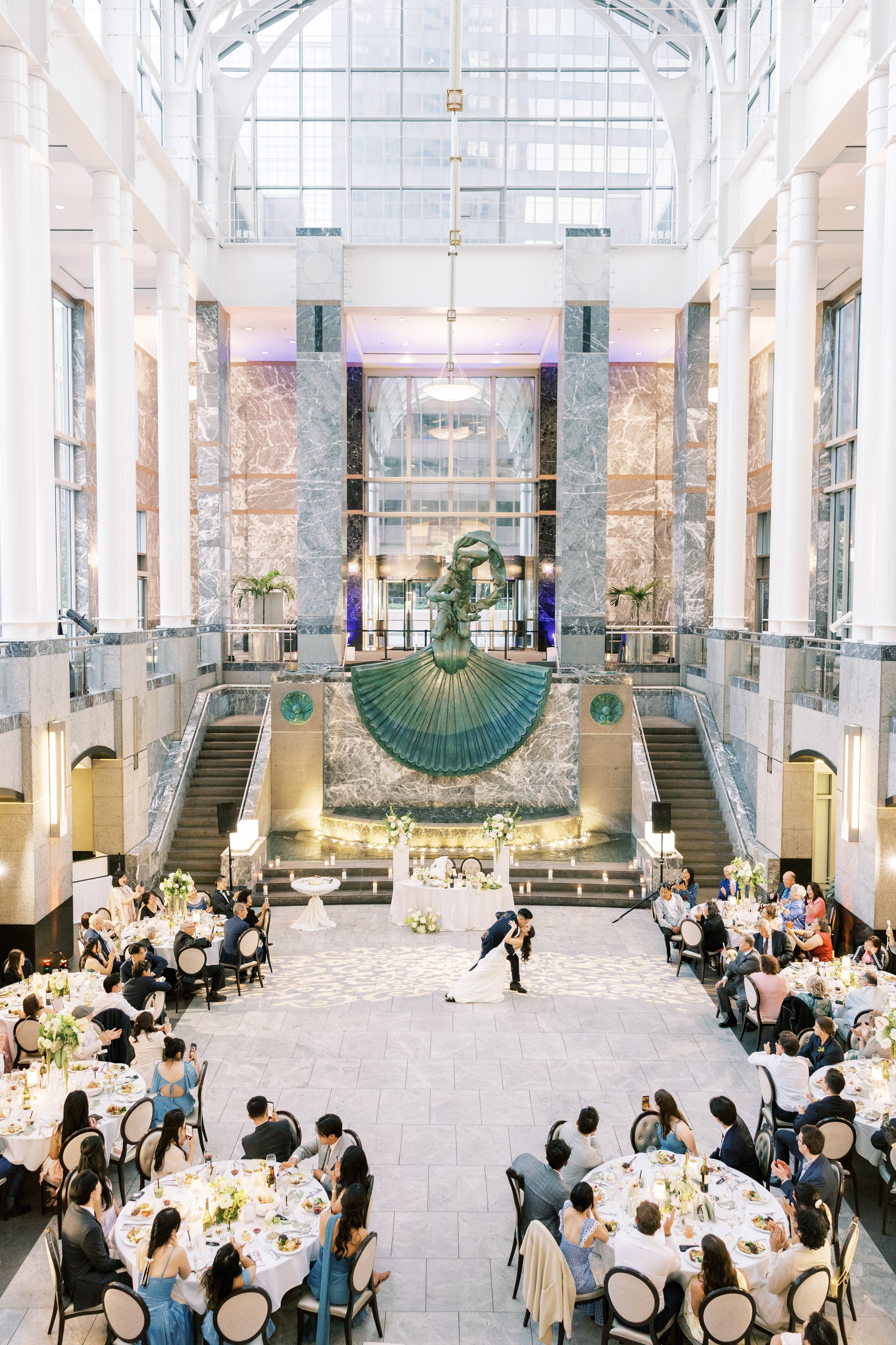 A wedding reception inside a grand atrium with a modern sculpture of a woman in a fan-shaped dress at the center. The bride and groom are dancing near the sculpture, surrounded by guest tables with formal dining settings.