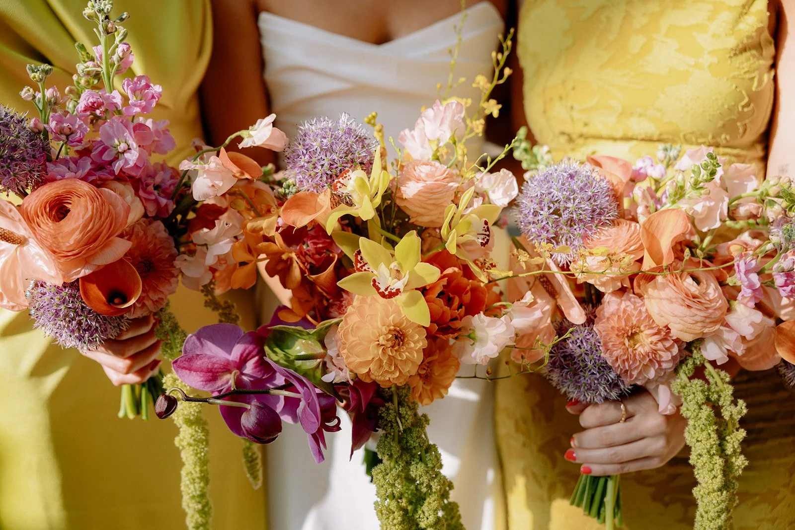 Three women holding large colorful bouquets of mixed flowers, including pink, orange, purple, and yellow blooms, at an event or celebration.