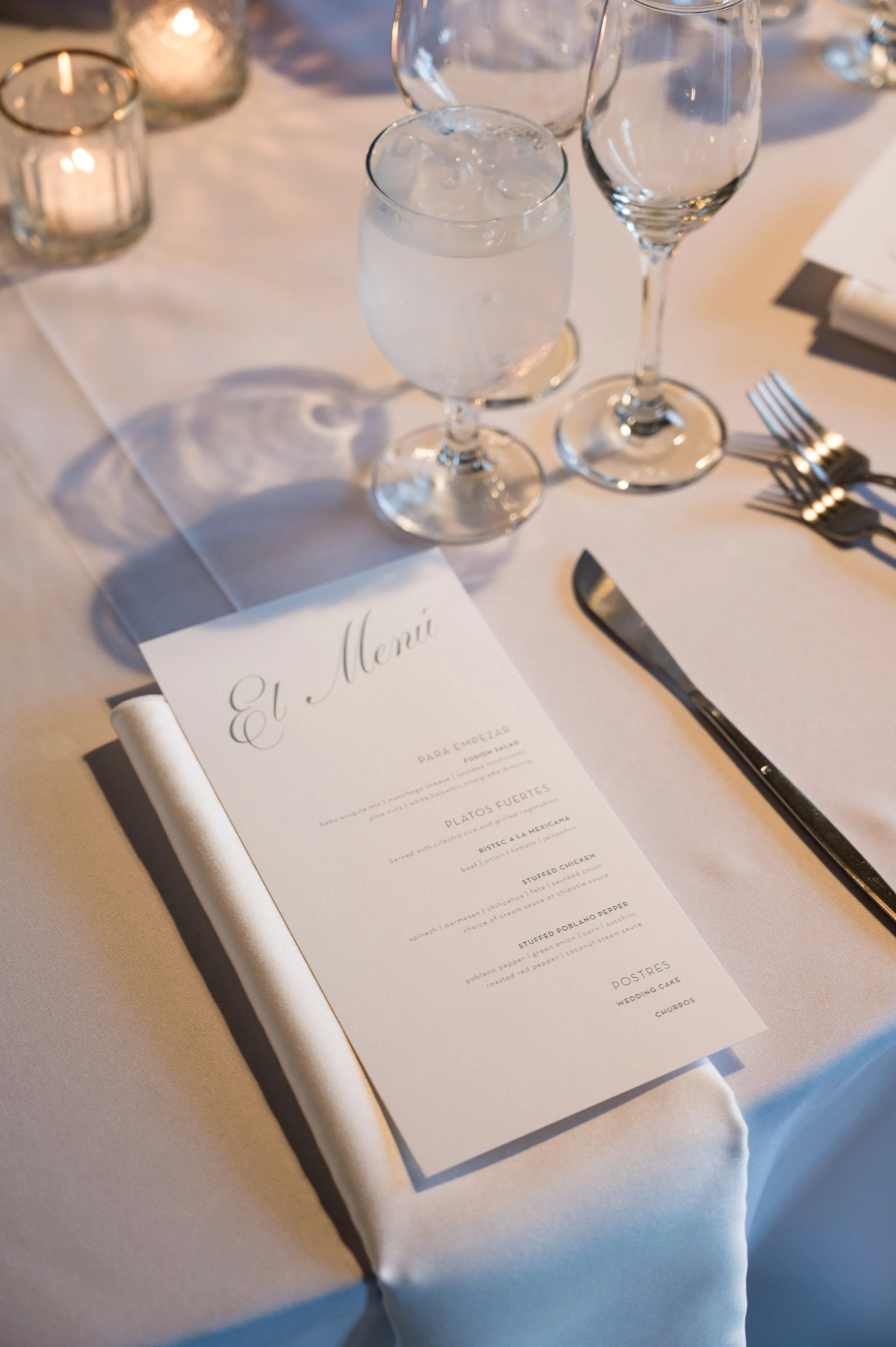 Elegant table setting with a wedding menu card, three empty champagne flutes, a water glass with ice, a set of three silver forks, and lit candles in small glass holders on a white tablecloth.