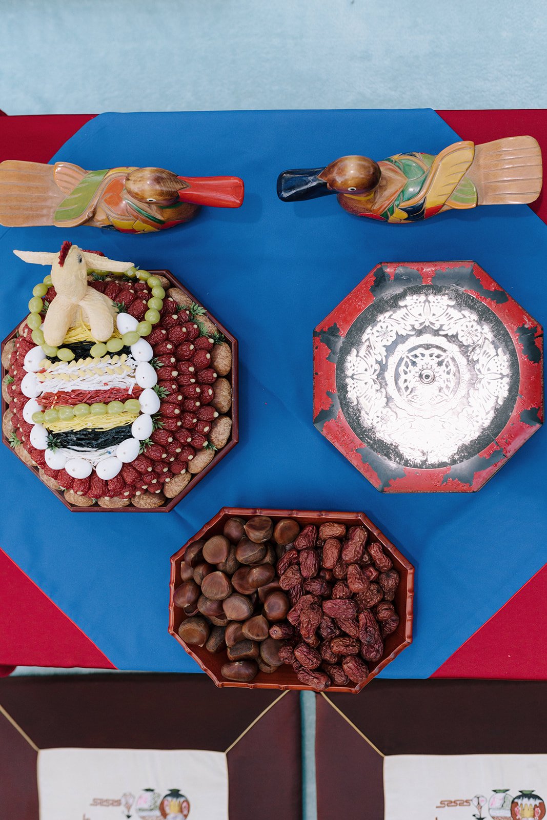 Decorative table display featuring carved wooden ducks, a fruit cake with marshmallow decorations, and bowls of chestnuts and dried red berries on a blue and red tablecloth.