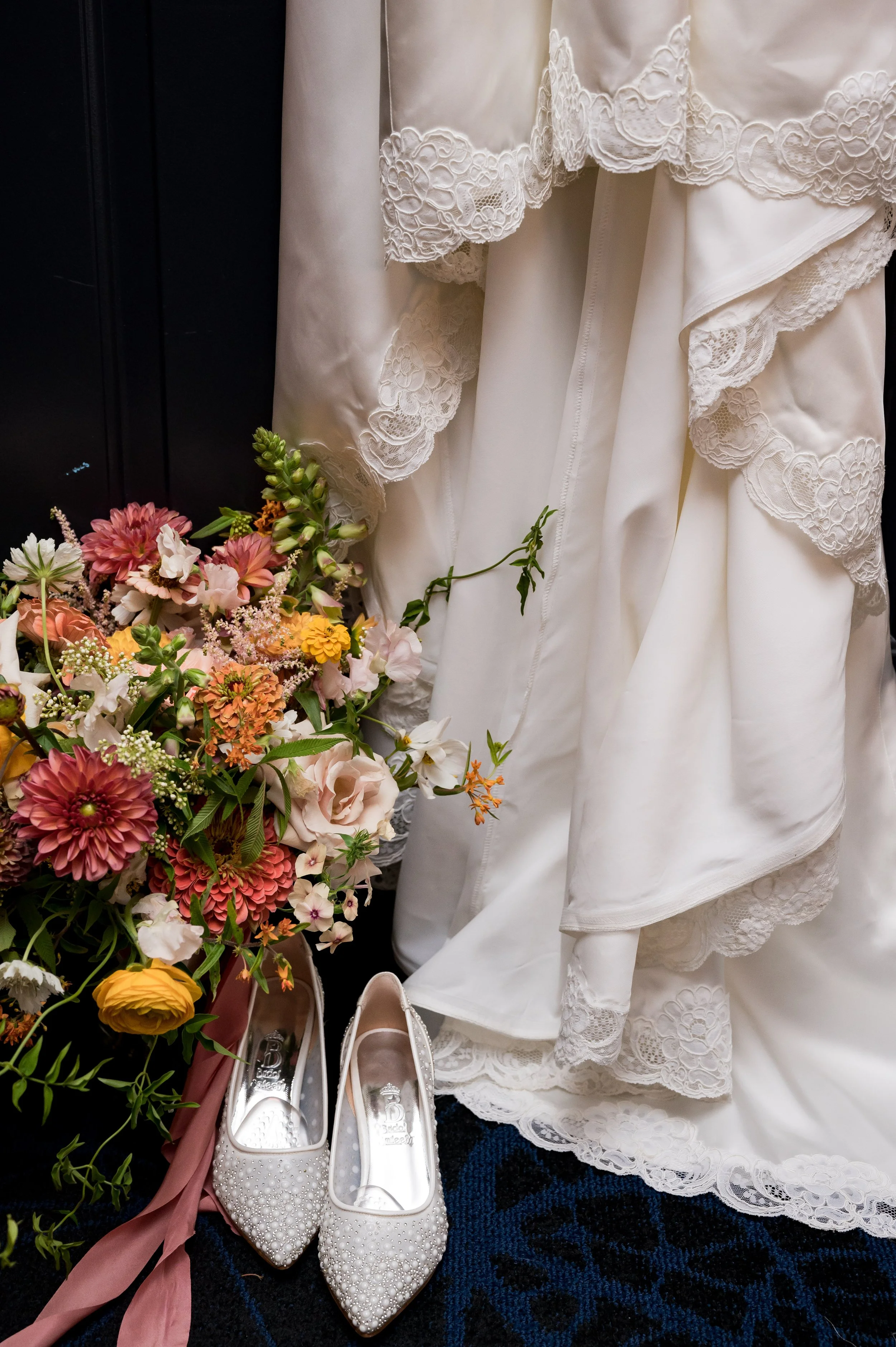 A collection of white bridal shoes decorated with pearls, a white wedding dress with lace details, and a bouquet of pink, yellow, and white flowers, all set against a dark background.