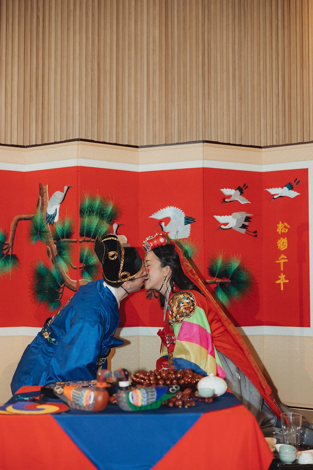 A couple dressed in traditional Chinese clothing sharing a kiss at a table with food and decorative items, with a red background featuring painted cranes and pine trees.