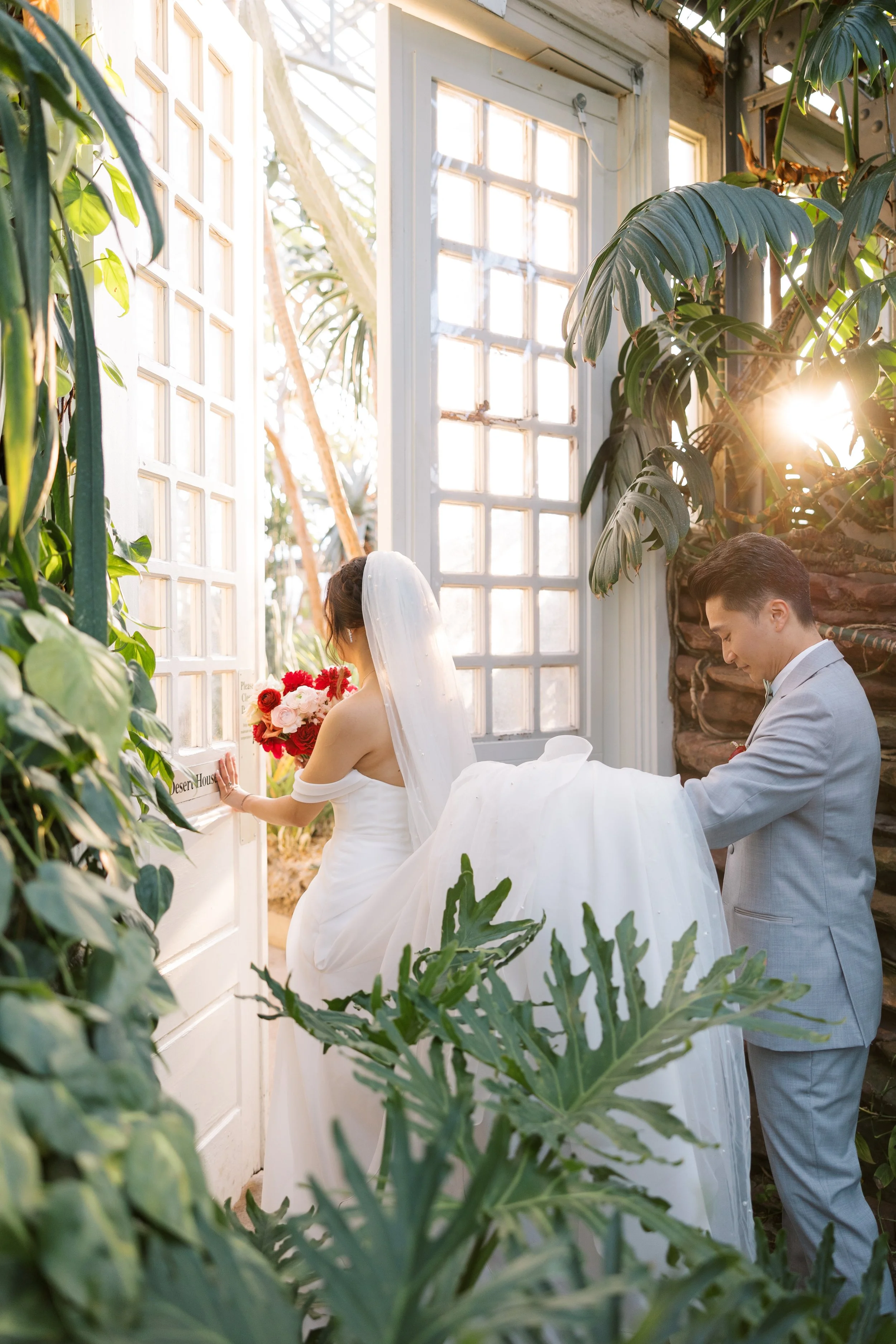 A bride and groom in wedding attire are inside a glass greenhouse, with lush green plants surrounding them and sunlight streaming through the windows.