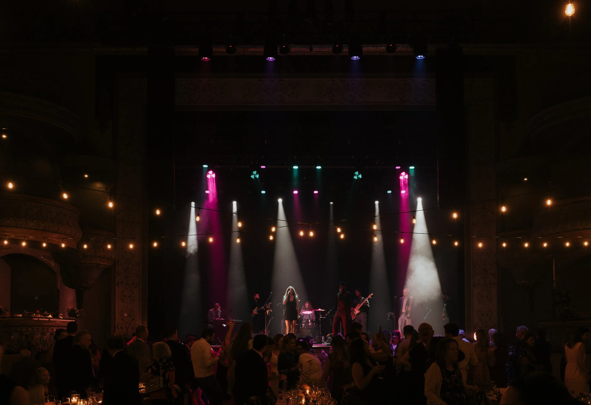 Concert performance on a stage with colorful lighting, band members playing instruments, audience seated at tables, ornate theater interior.