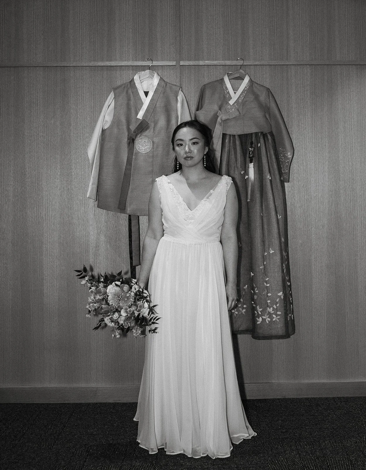 A woman in a white wedding dress holding a bouquet stands in front of traditional Korean clothing hanging on a wall.