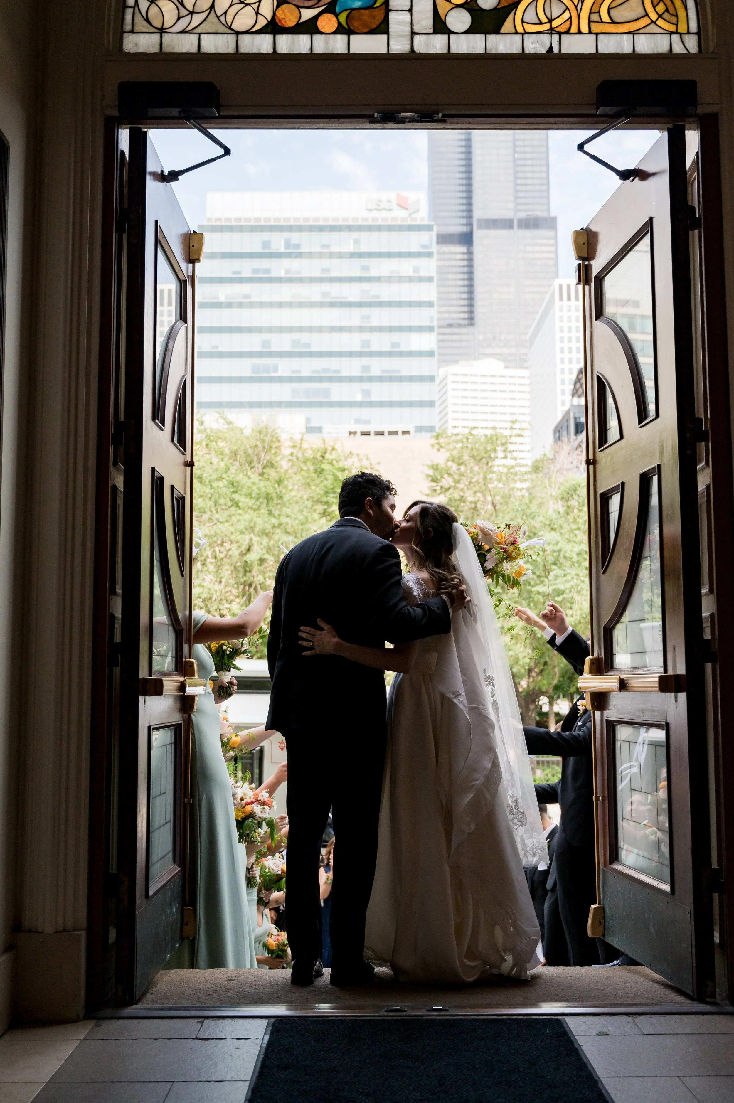 A bride and groom share a kiss at their wedding ceremony, seen through open church doors with a cityscape background.