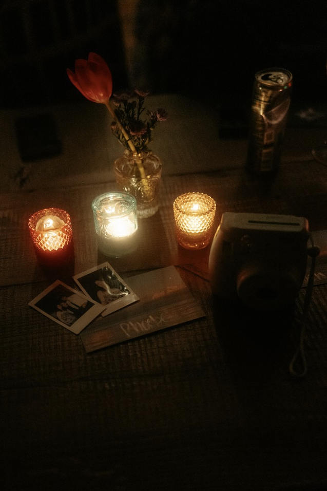 A wooden table with four lit candles in glass holders, a small glass jar with a flower, Polaroid photos, a menu, a camera, and a can in the background.