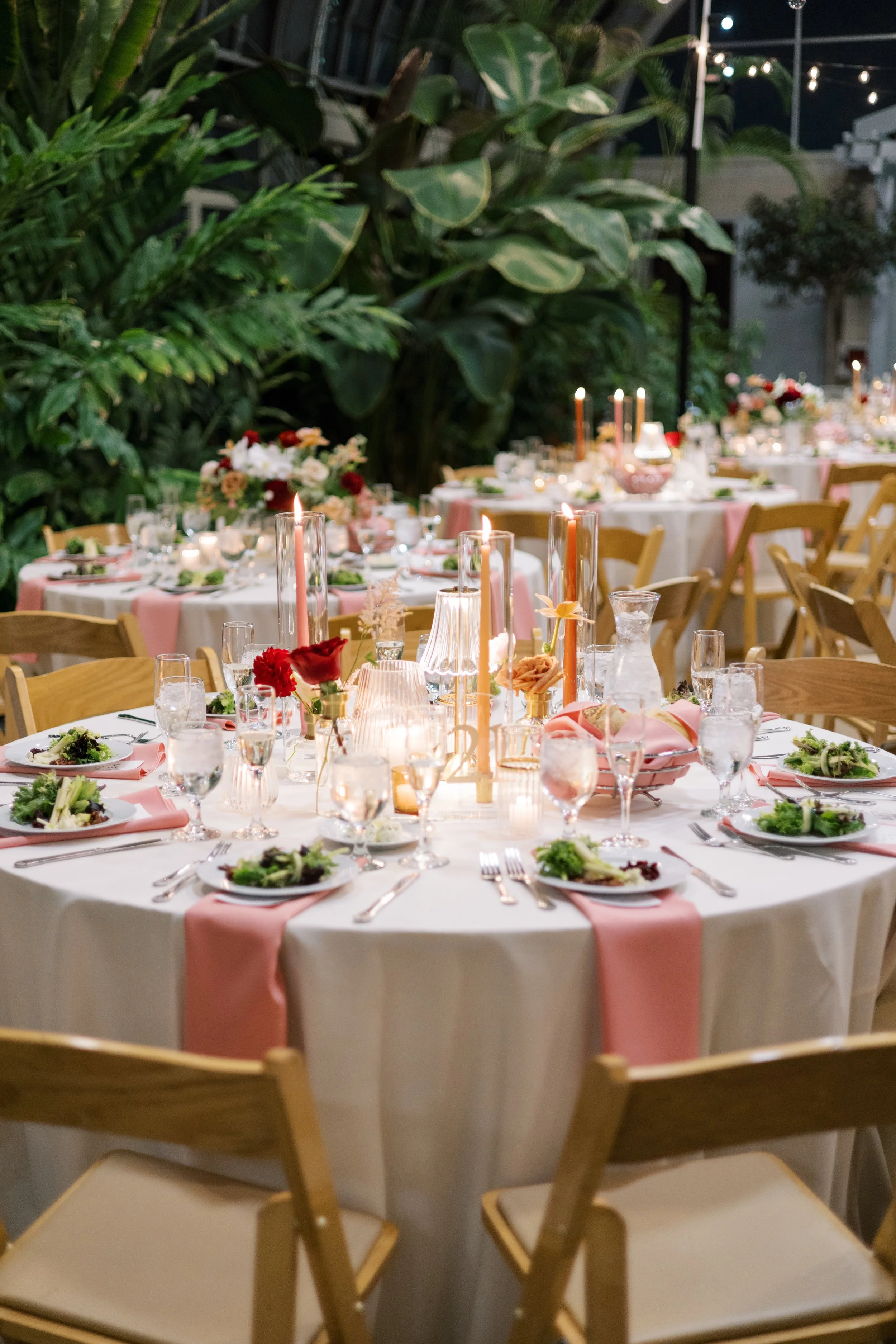 Elegant outdoor dining setup with round tables covered in white tablecloths, decorated with pink runners, floral centerpieces, and tall pink candles, surrounded by wooden chairs, greenery, and string lights in the background.