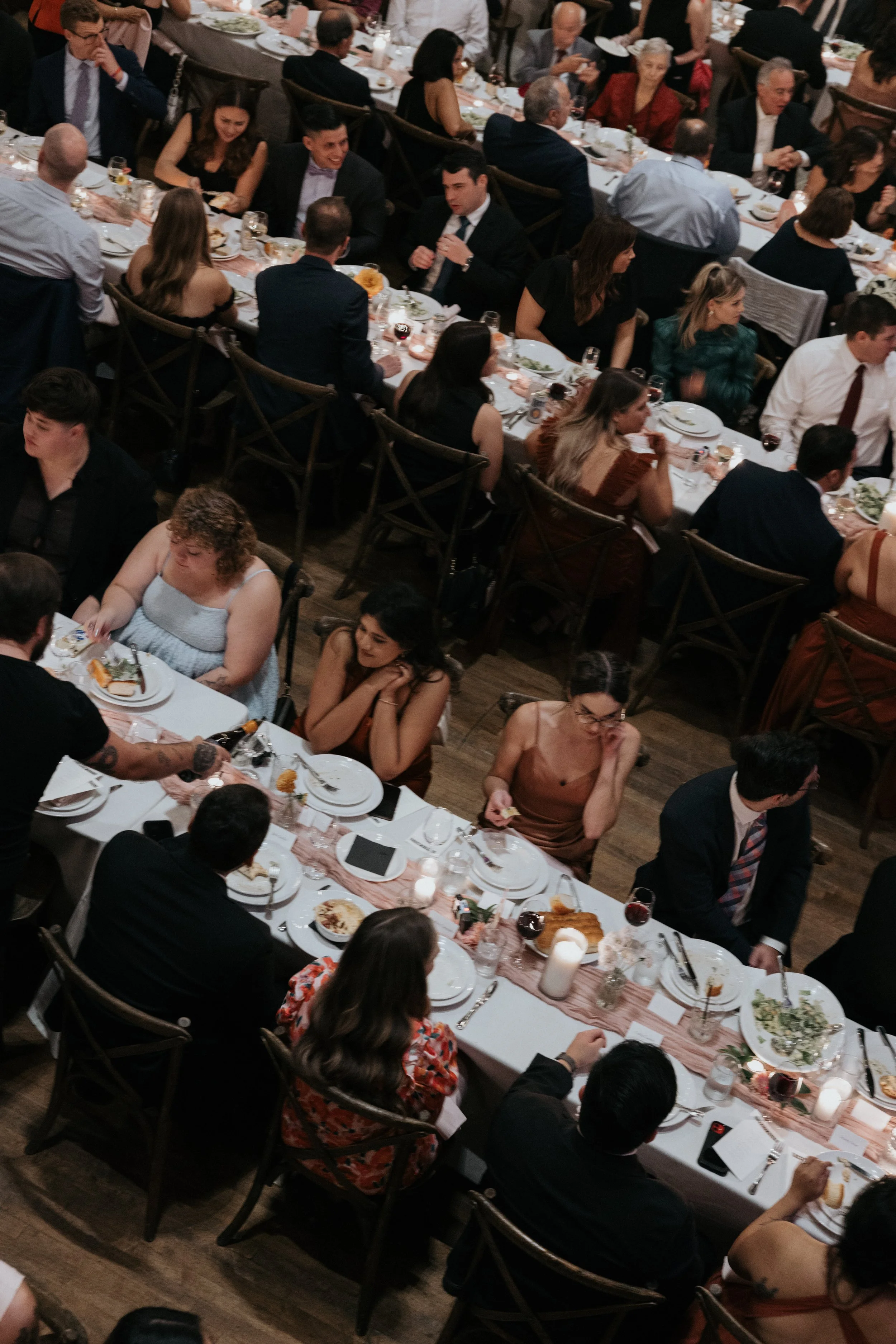 A large formal dinner party with many guests seated at long tables filled with plates of food, glasses of wine, and candles in a dimly lit room.