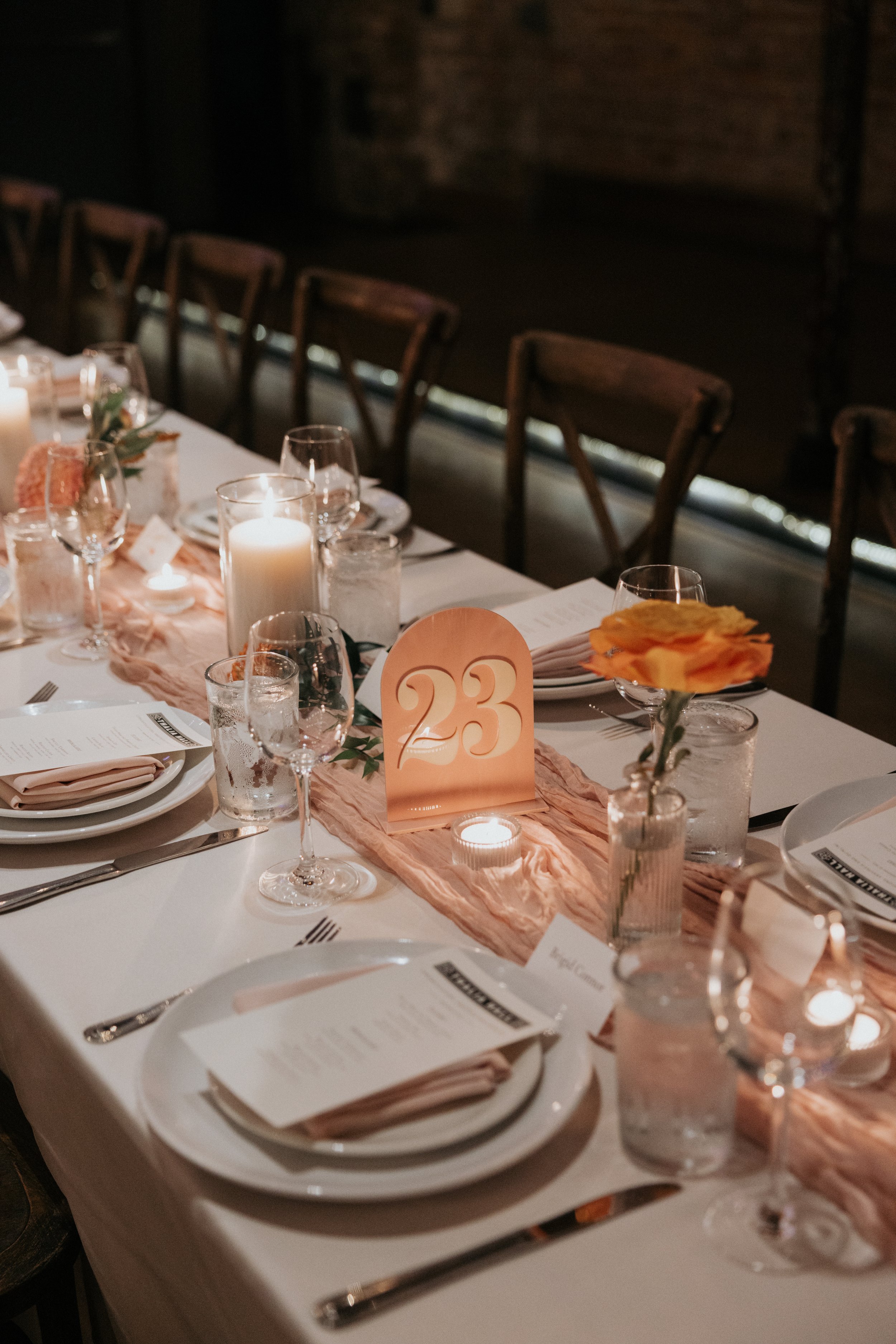 A formal dining table set with white tablecloth, plates, wine glasses, water glasses, silverware, and menu cards. The table is decorated with candles in glass holders, floral arrangements, and a pink table runner. There is a prominent table number '2
