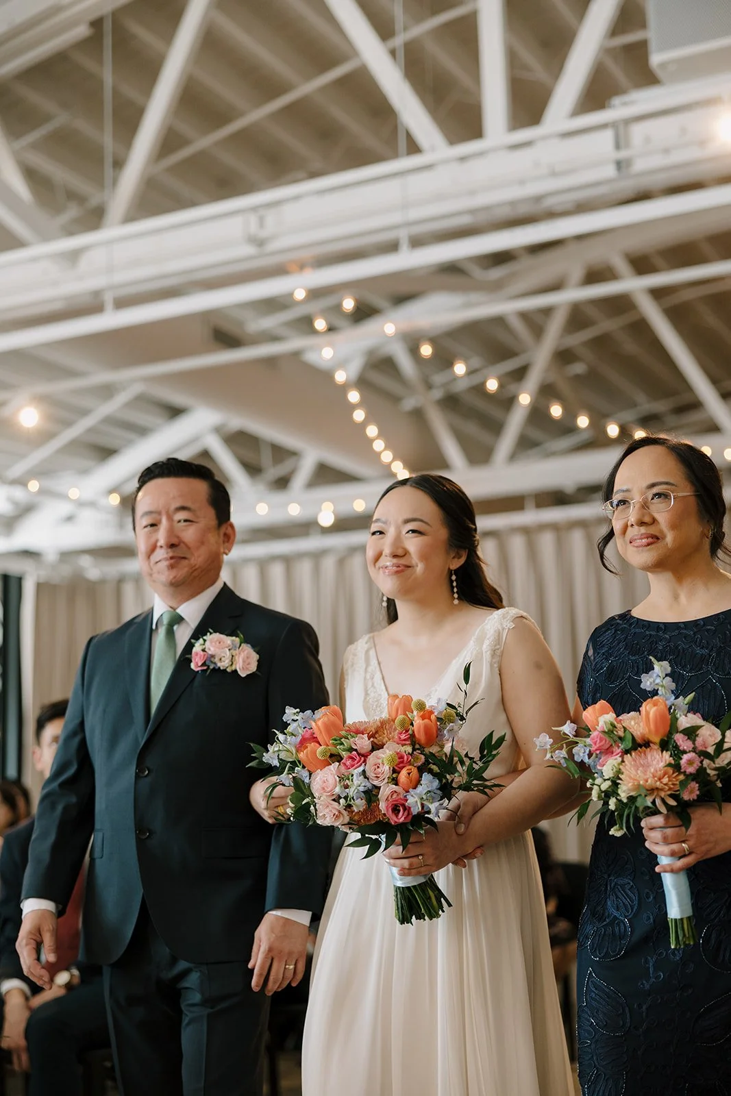 A bride in a wedding dress walking down the aisle with her parents at her side, holding a bouquet of flowers, in a decorated wedding venue with string lights overhead.