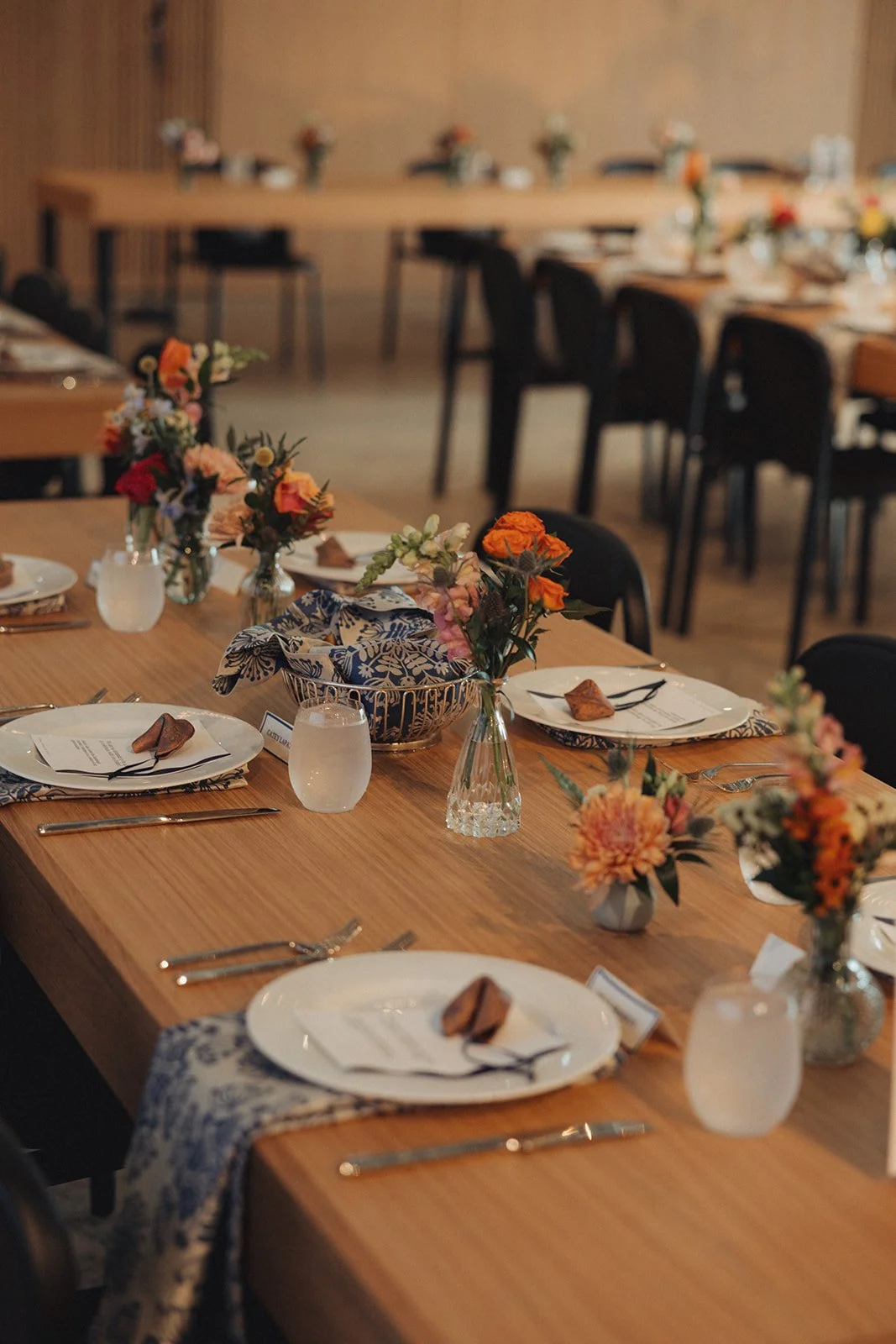 Decorated dining table with floral centerpieces, plates, napkins, cutlery, and glasses in a warmly lit room, prepared for a meal or event.