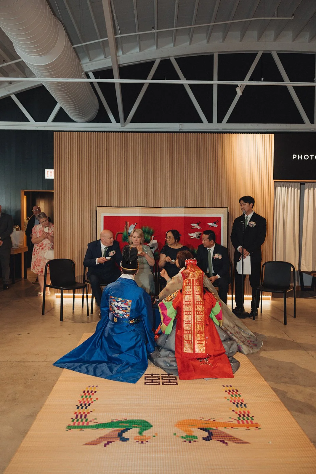 Traditional Korean wedding ceremony with a bride and groom in hanbok, sitting on a woven mat, as elderly guests seated in chairs observe. Bridesmaids, groomsmen, and other guests stand nearby in a decorated indoor setting.