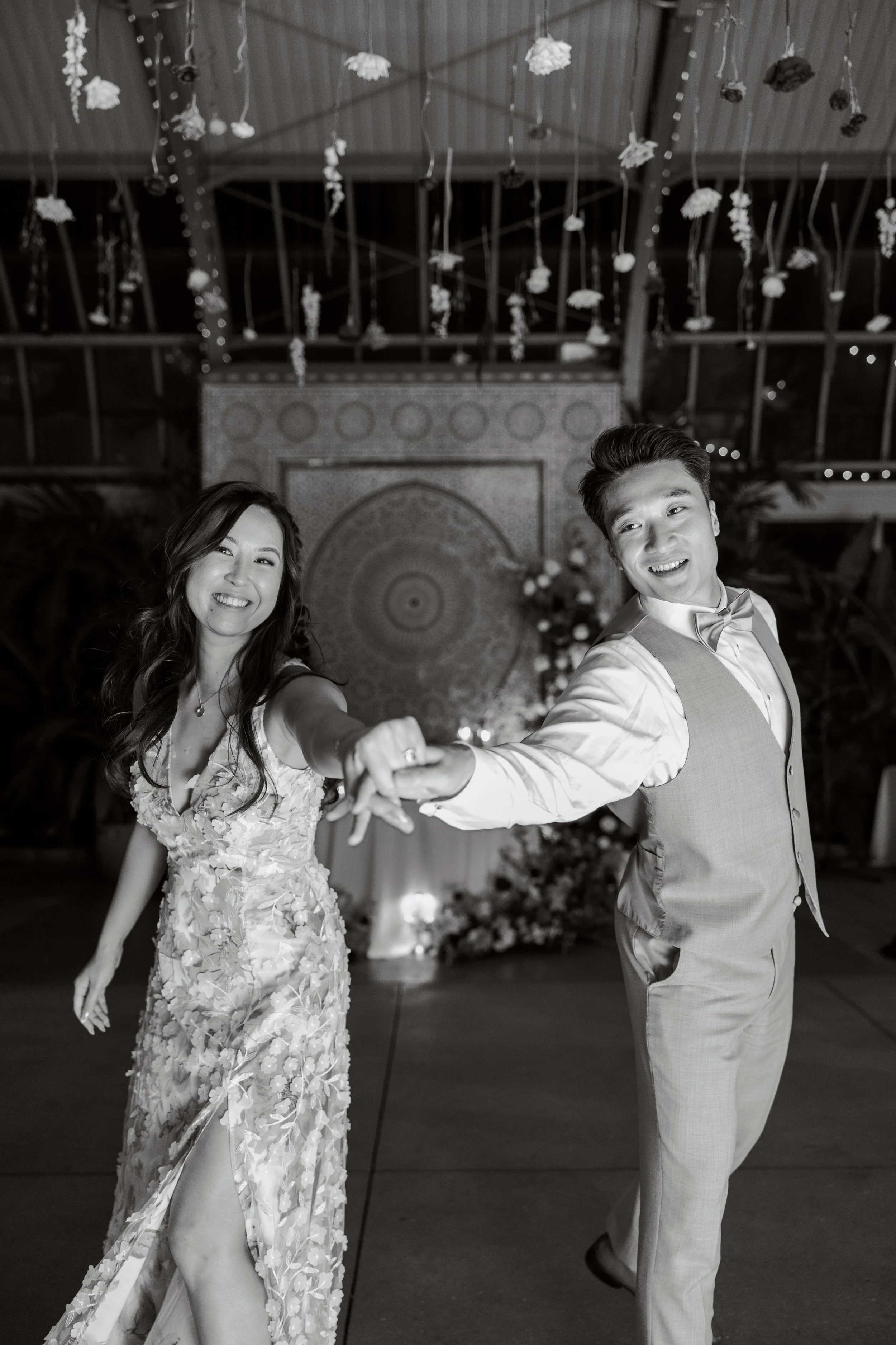A bride and groom dancing at their wedding reception, holding hands and smiling. The bride wears a floral lace wedding dress with a slit, and the groom is in a light-colored suit with a bow tie. The background is decorated with hanging flowers and st