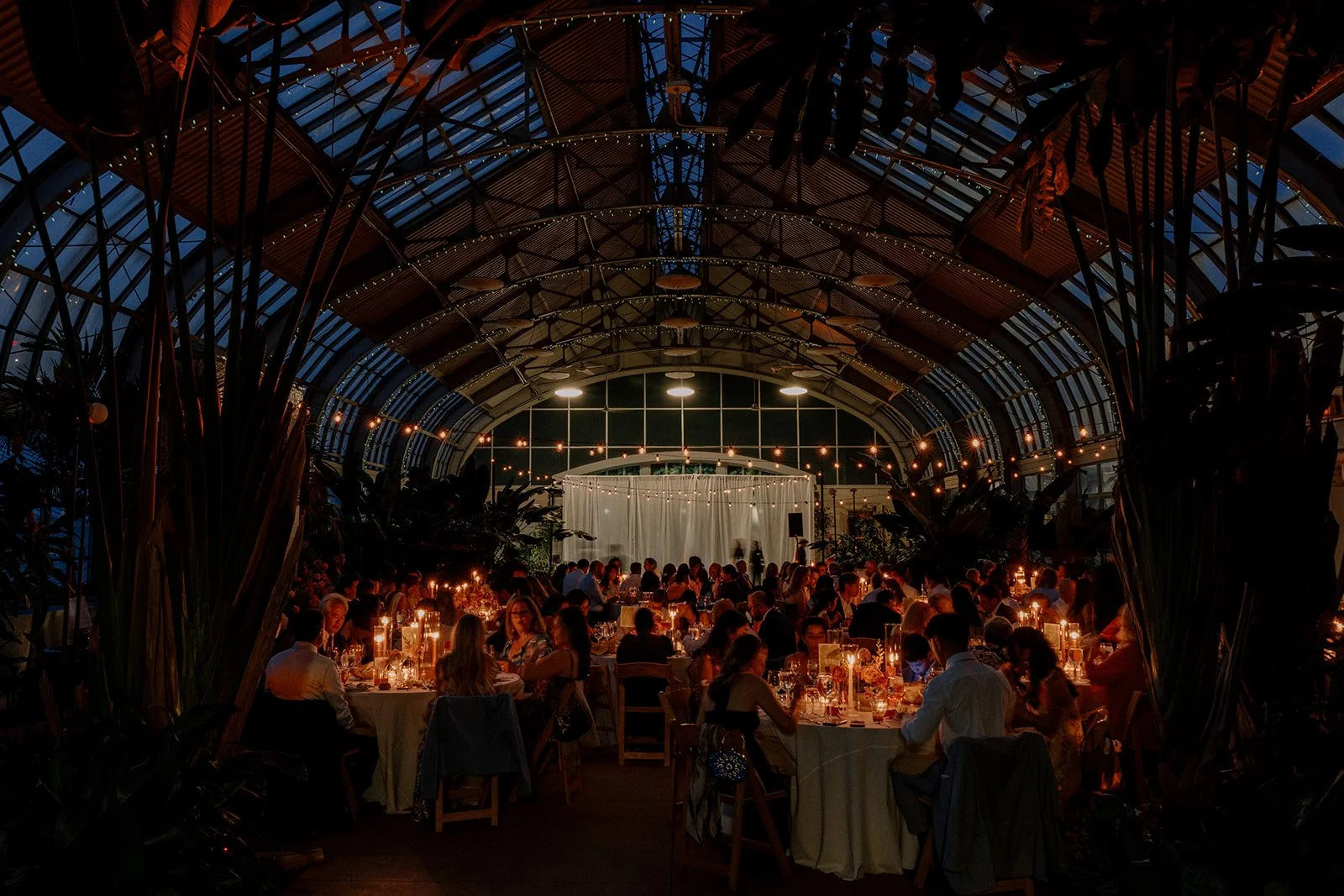 Indoor event space filled with people seated at decorated tables, illuminated by string lights and candles, with a glass-domed roof and a white curtain backdrop.