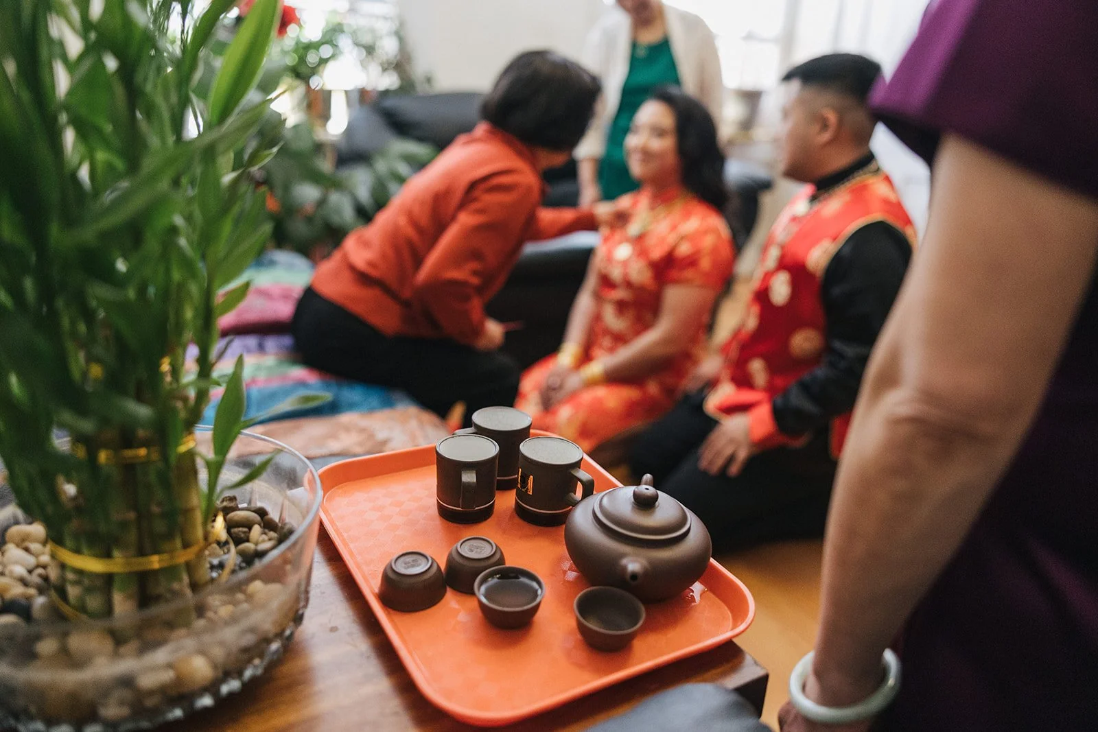Tea set with cups and teapot on a tray, in front of a group of people celebrating an occasion with traditional attire, while a green plant is visible in the foreground.