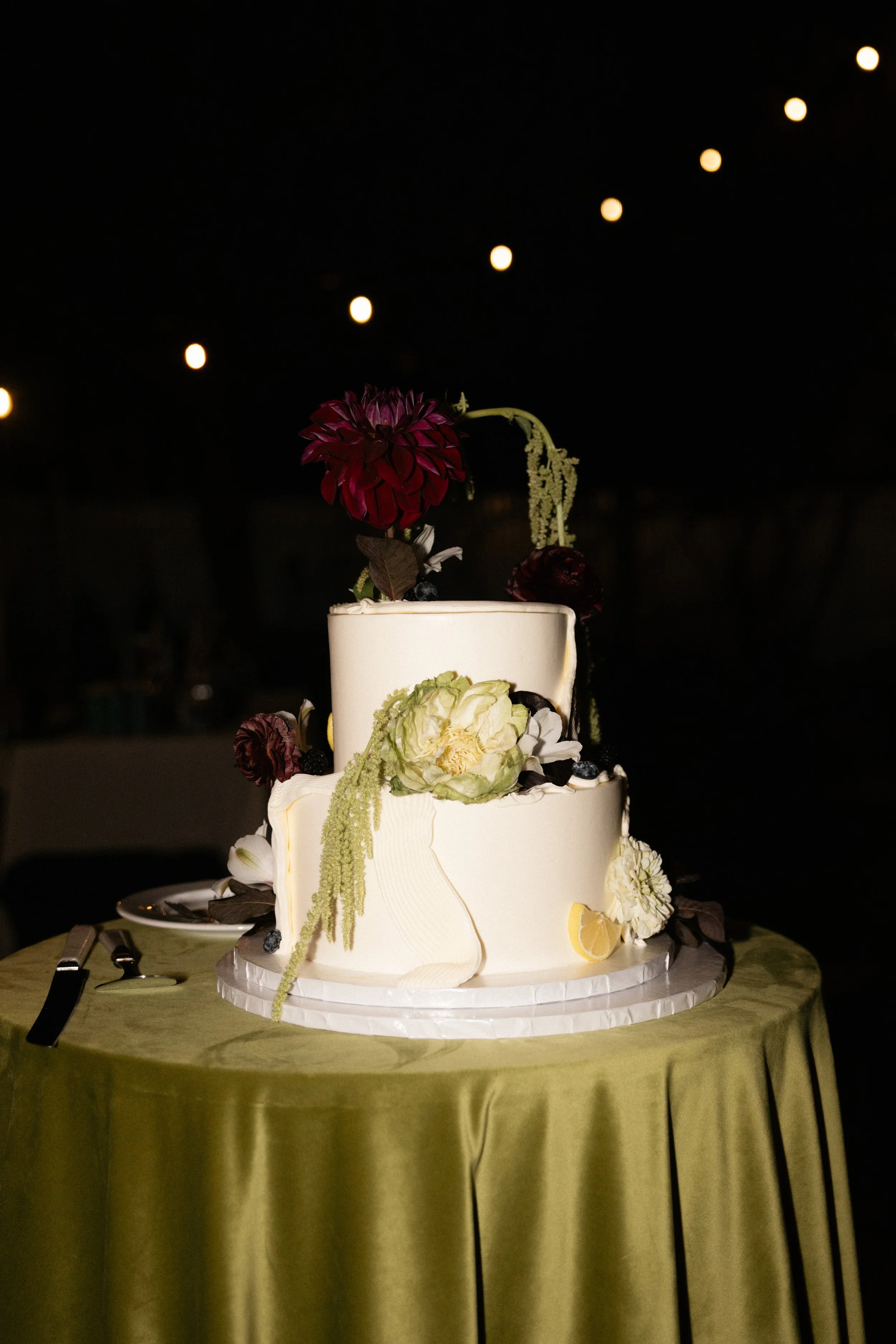 A tiered white wedding cake decorated with flowers and lemons, placed on a green tablecloth, with a dark background and string lights overhead.