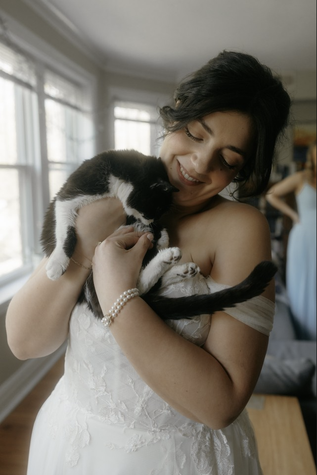 A woman in a white wedding dress holding a black and white cat indoors near windows.