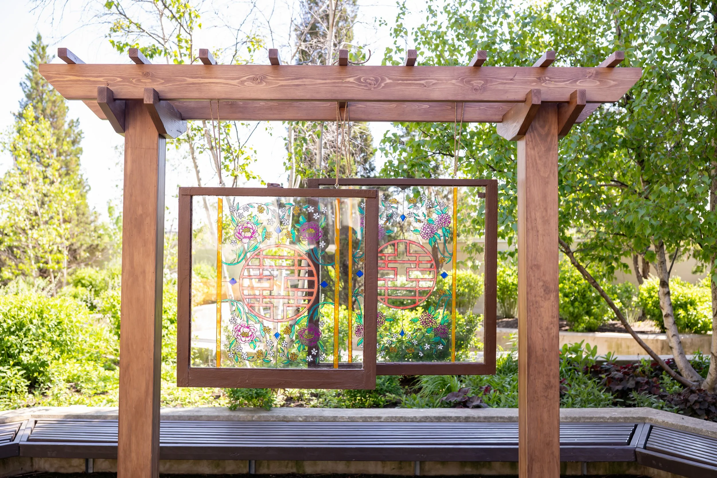 Wooden garden arbor with decorative glass panels featuring floral and geometric designs, set in a lush green garden with trees and shrubs in the background.