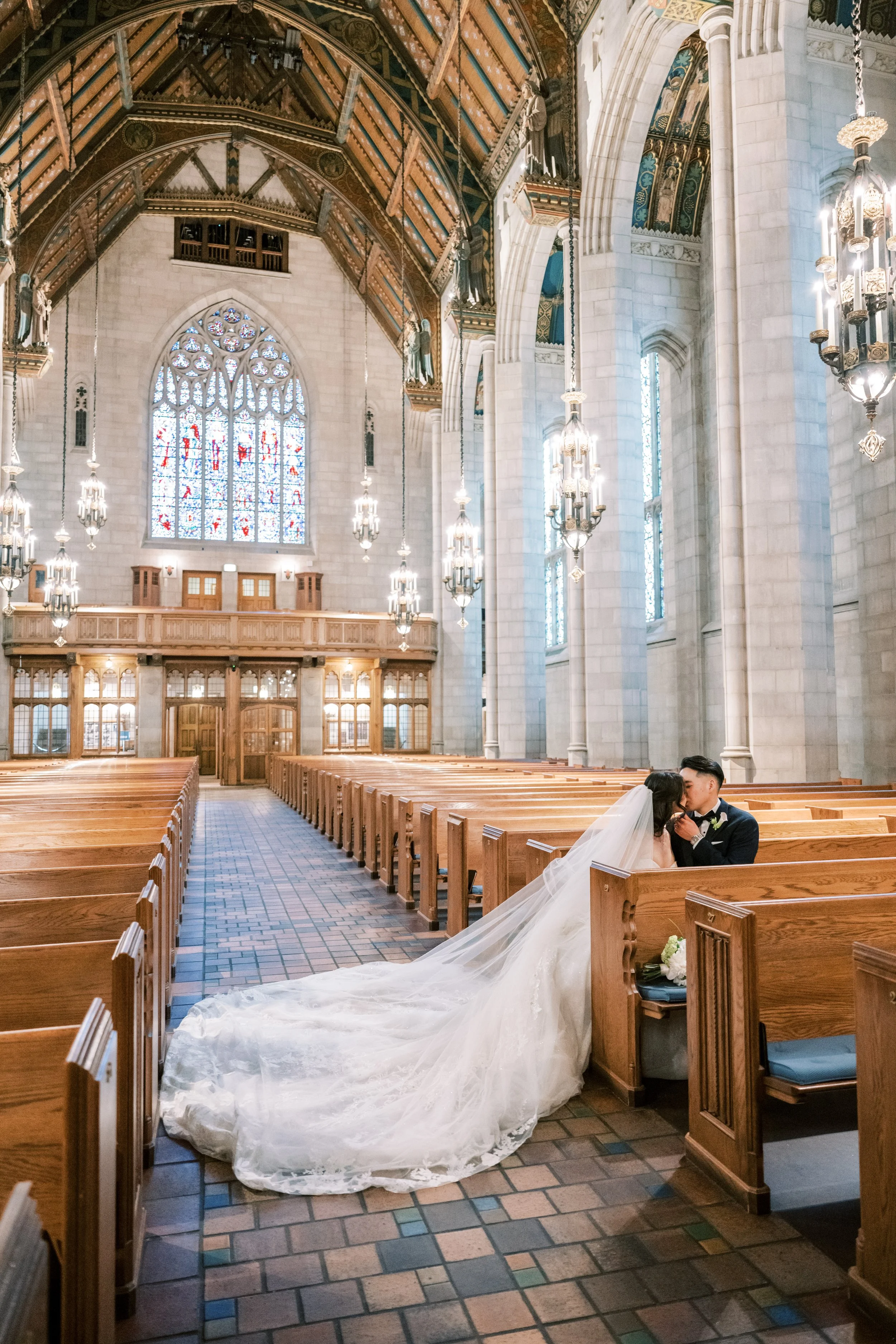 A bride and groom sitting closely and kissing in an empty church with wood pews, stained glass windows, and ornate hanging chandeliers.