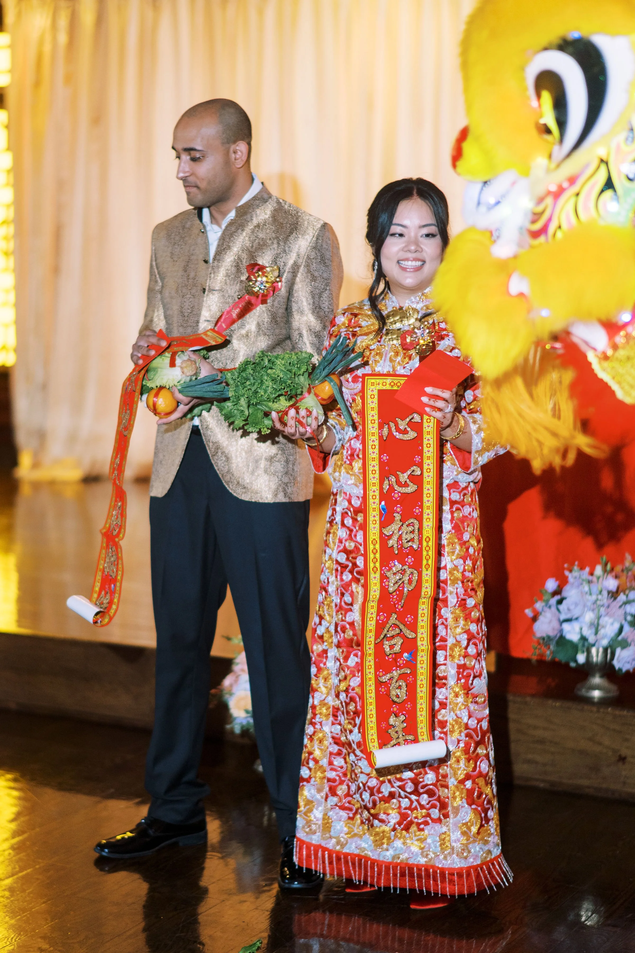 People participating in a traditional Chinese celebration or ceremony, with one holding a scroll and fruit, and another dressed in a golden and red embroidered robe surrounded by lion dance costume and floral decorations.