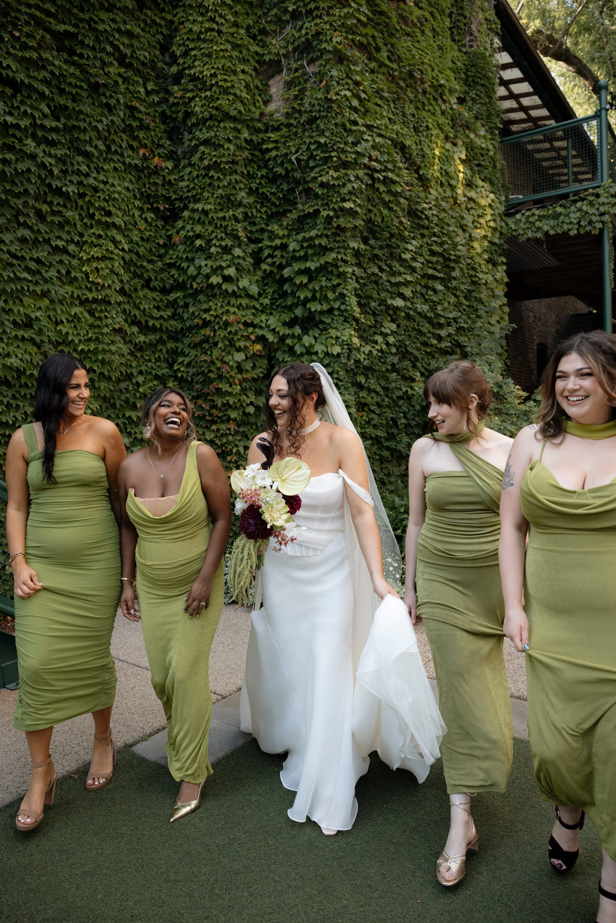 A bride and four bridesmaids walking together outside, laughing, and dressed in wedding attire. The bride is holding a bouquet of flowers and is wearing a white gown with a veil. The bridesmaids are dressed in matching green dresses and high heels. T