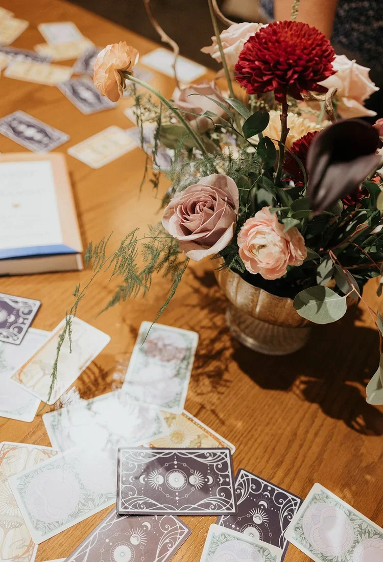 A floral centerpiece with various flowers including roses and dahlias on a wooden table surrounded by tarot or oracle cards.
